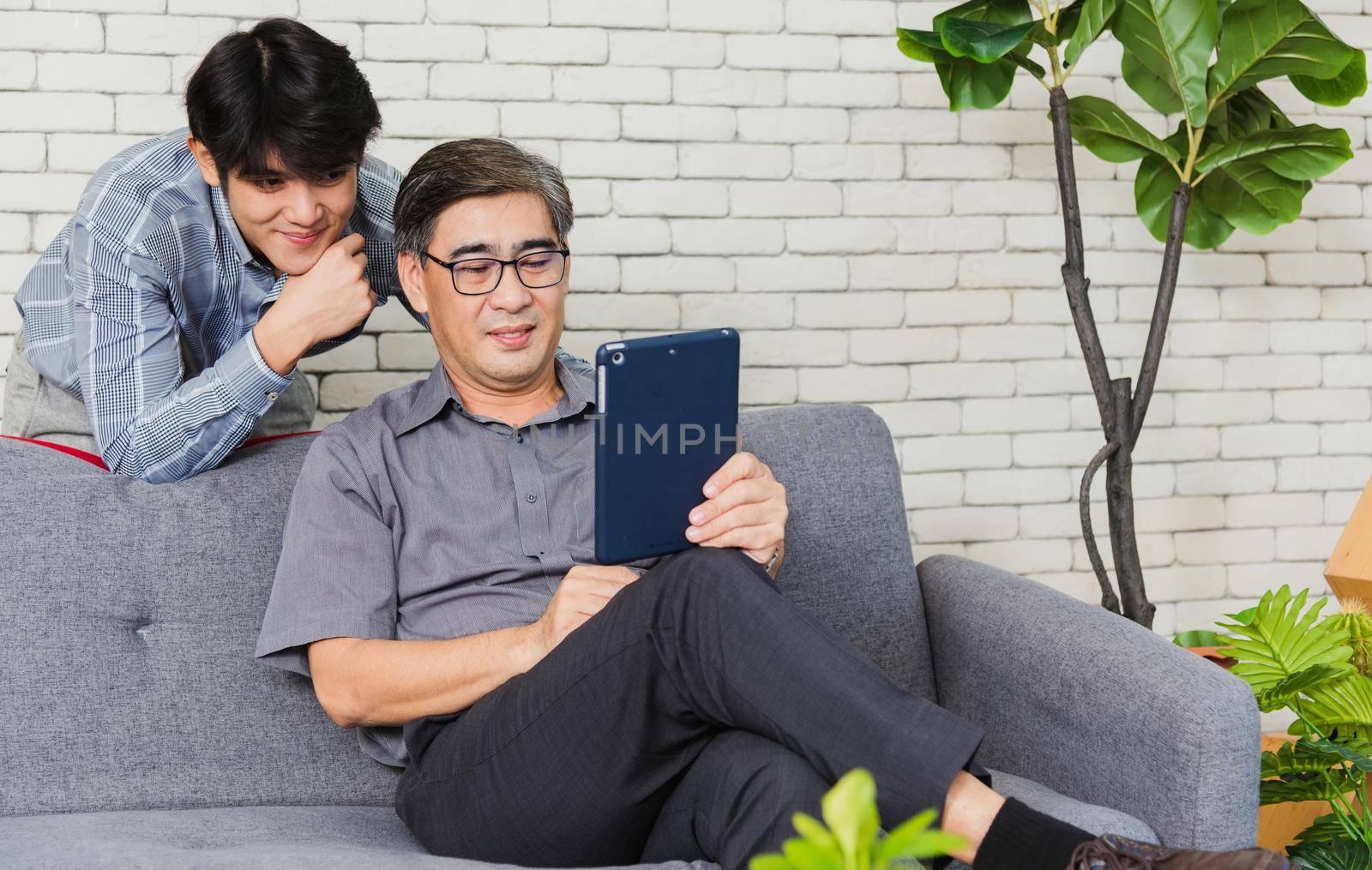 Asian senior businessman with digital tablet discuss together with young team in office. Father man and his son sit on sofa talking chatting on video call conference on tablet in living room at home