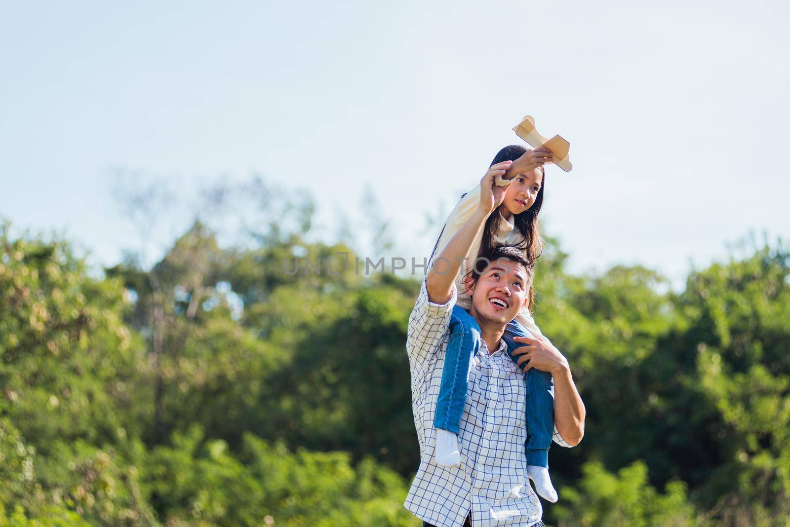 father and carrying an excited girl on shoulders having fun and enjoying outdoor lifestyle together playing aircraft toy by Sorapop