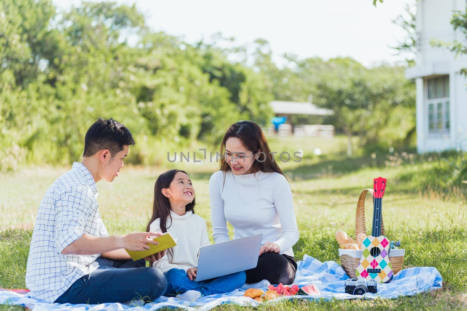 Happy Asian lifestyle young family father, mother and little girl having fun outdoor sitting on picnic blanket using laptop computer technologies while relaxing to open song at garden park in summer