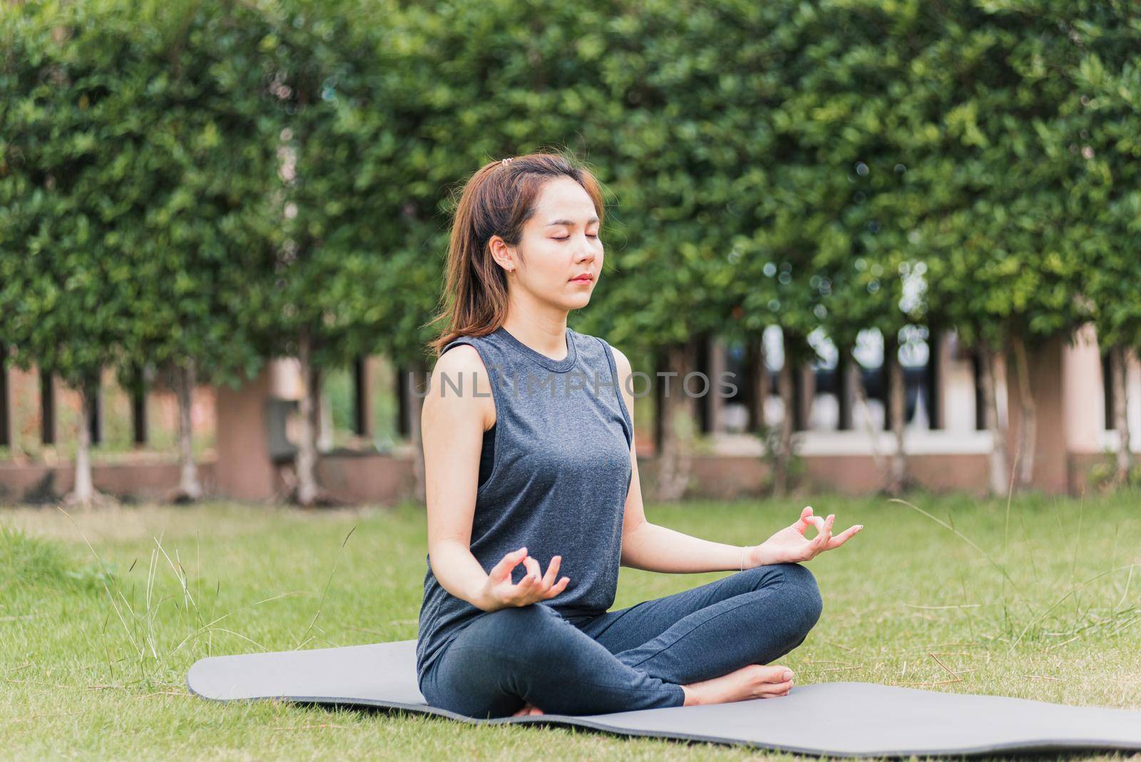 Asian young woman doing yoga outdoors in meditate lotus pose sitting on green grass with closed eyes at the garden park, health care concept