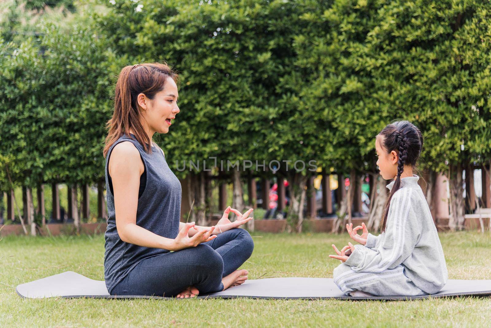 mother practicing doing yoga exercises with her daughter outdoors in meditate pose together by Sorapop