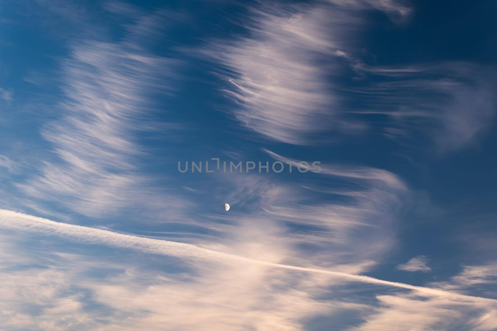 Cirrus clouds against a blue, dark blue sky, a small moon among the clouds. Airplane trail in the sky.