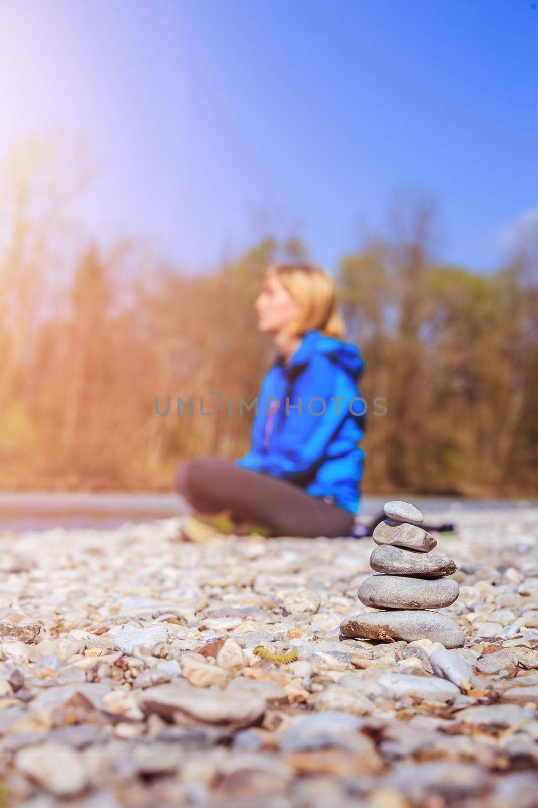 Cairn on a pebble beach, meditating woman in the background. Morning sun.