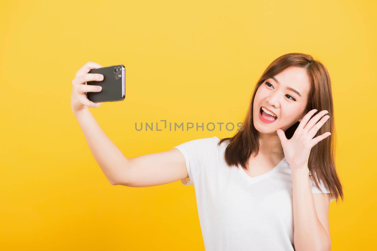 Asian happy portrait beautiful cute young woman teen smiling standing making selfie photo, video call on smartphone raise hand to say hello isolated, studio shot yellow background with copy space