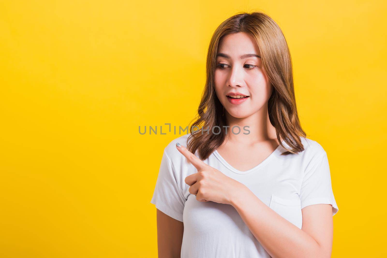 Asian Thai happy portrait beautiful cute young woman standing wear white t-shirt pointing finger away side looking to away side, studio shot isolated on yellow background with copy space