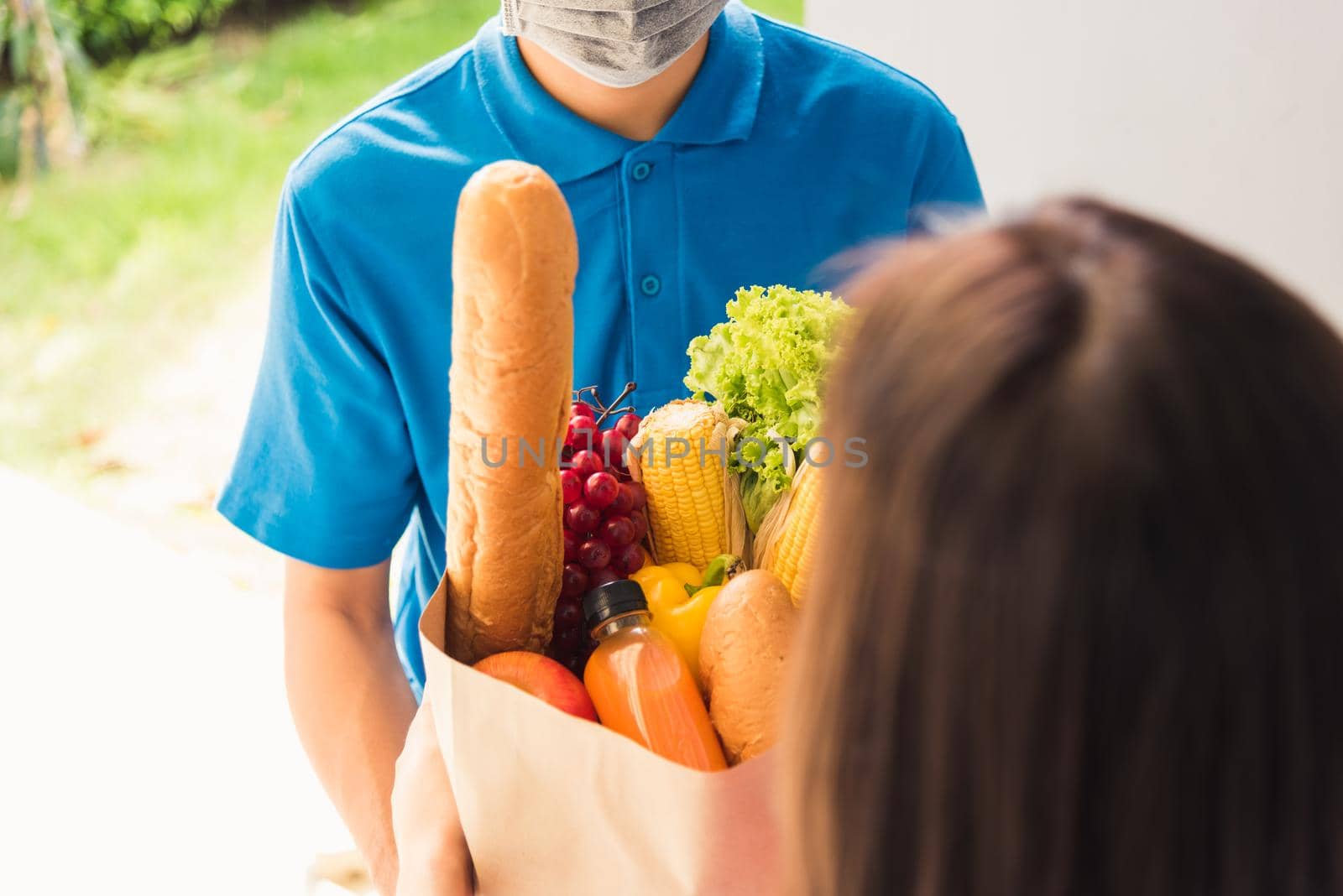 Asian young delivery man in uniform wear protective face mask he making grocery service giving fresh food to woman customer receiving front house under pandemic coronavirus, Back to new normal concept