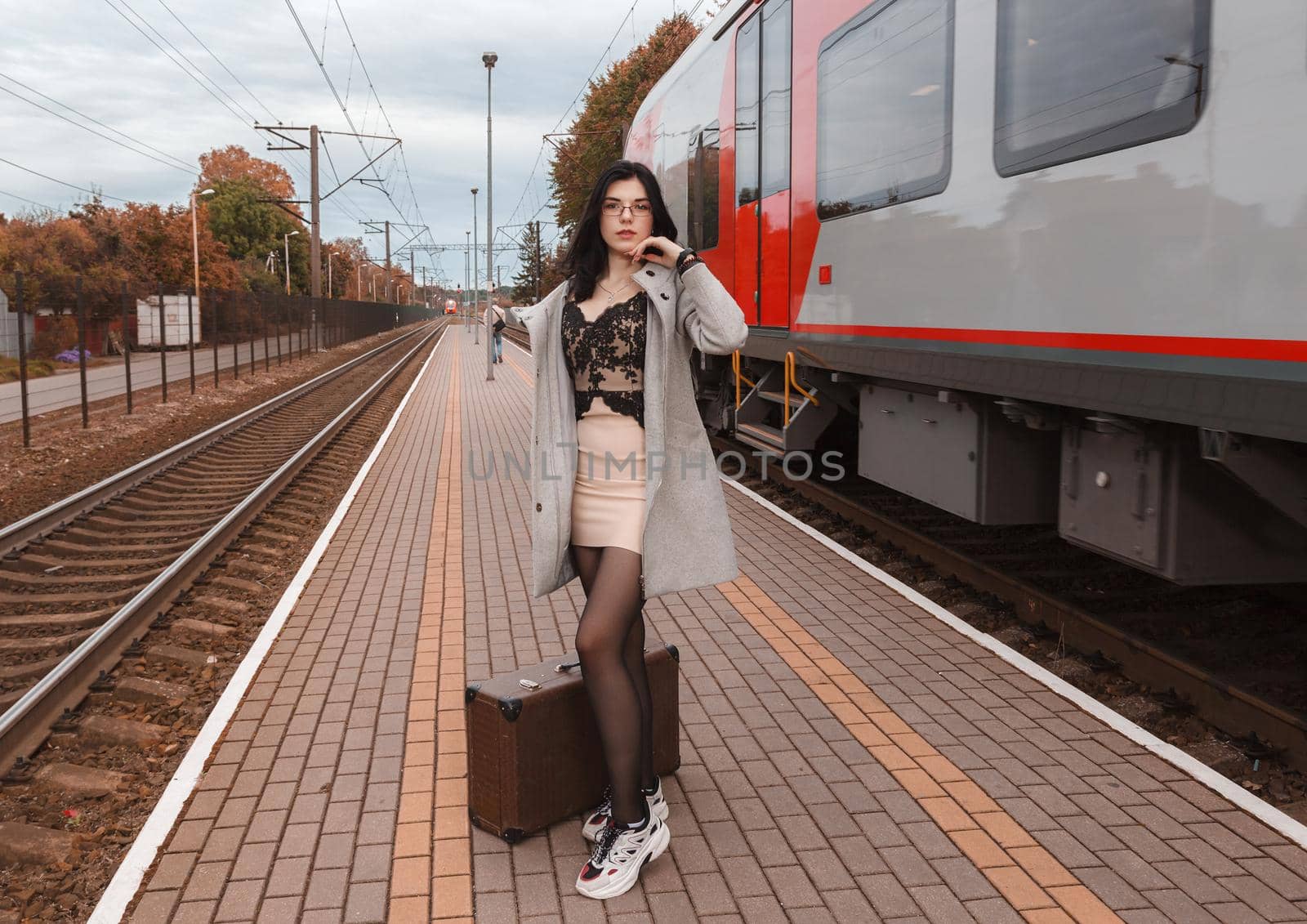 lonely young girl in gray coat with suitcase standing at railway station on gloomy autumn day
