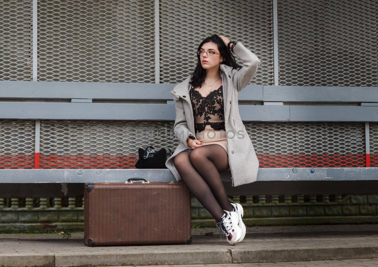 lonely young girl in a gray coat with a suitcase sitting on a bench at a railway station in the late evening