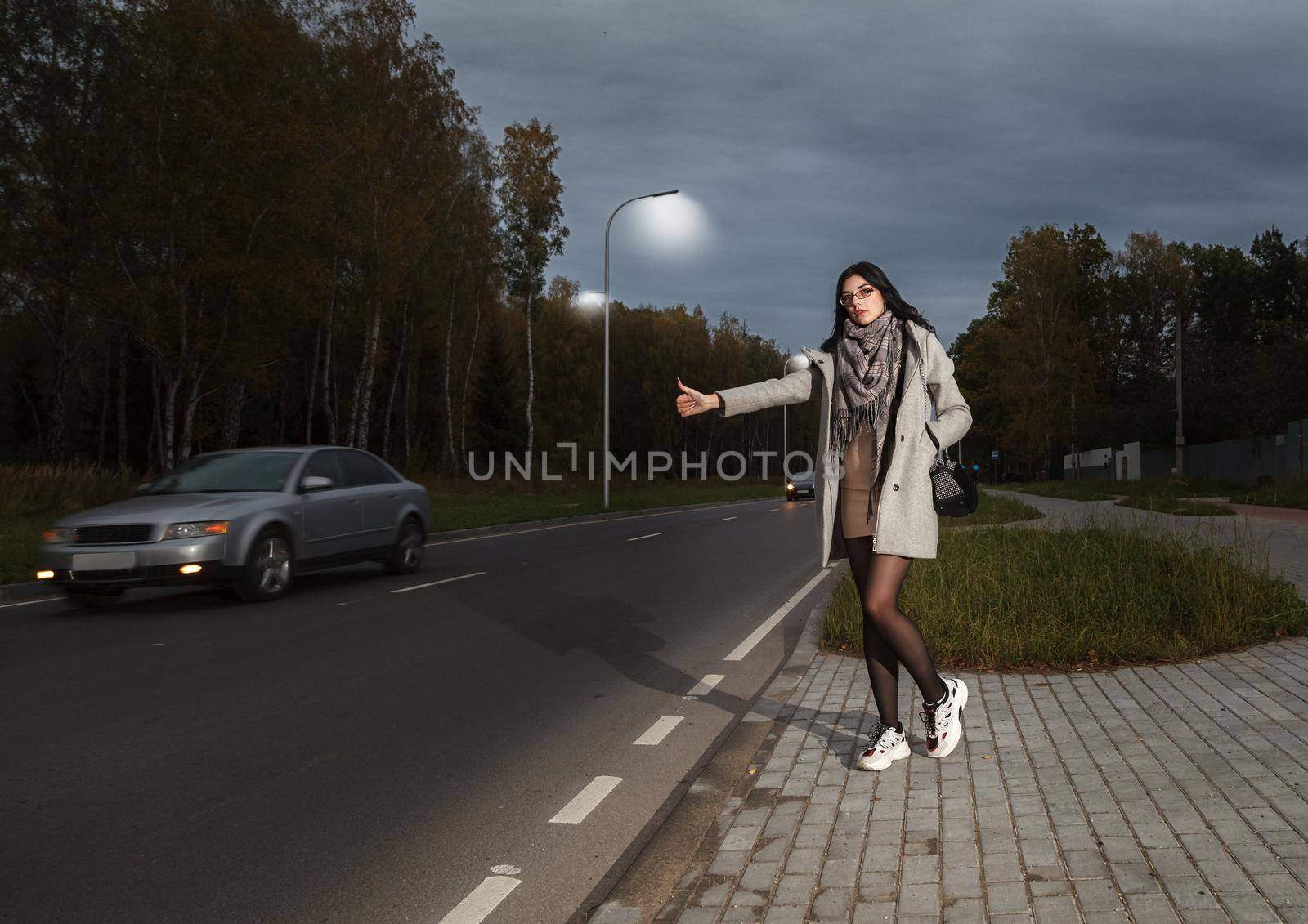 young girl in a gray coat stands near the road trying to stop a car in a late autumn evening