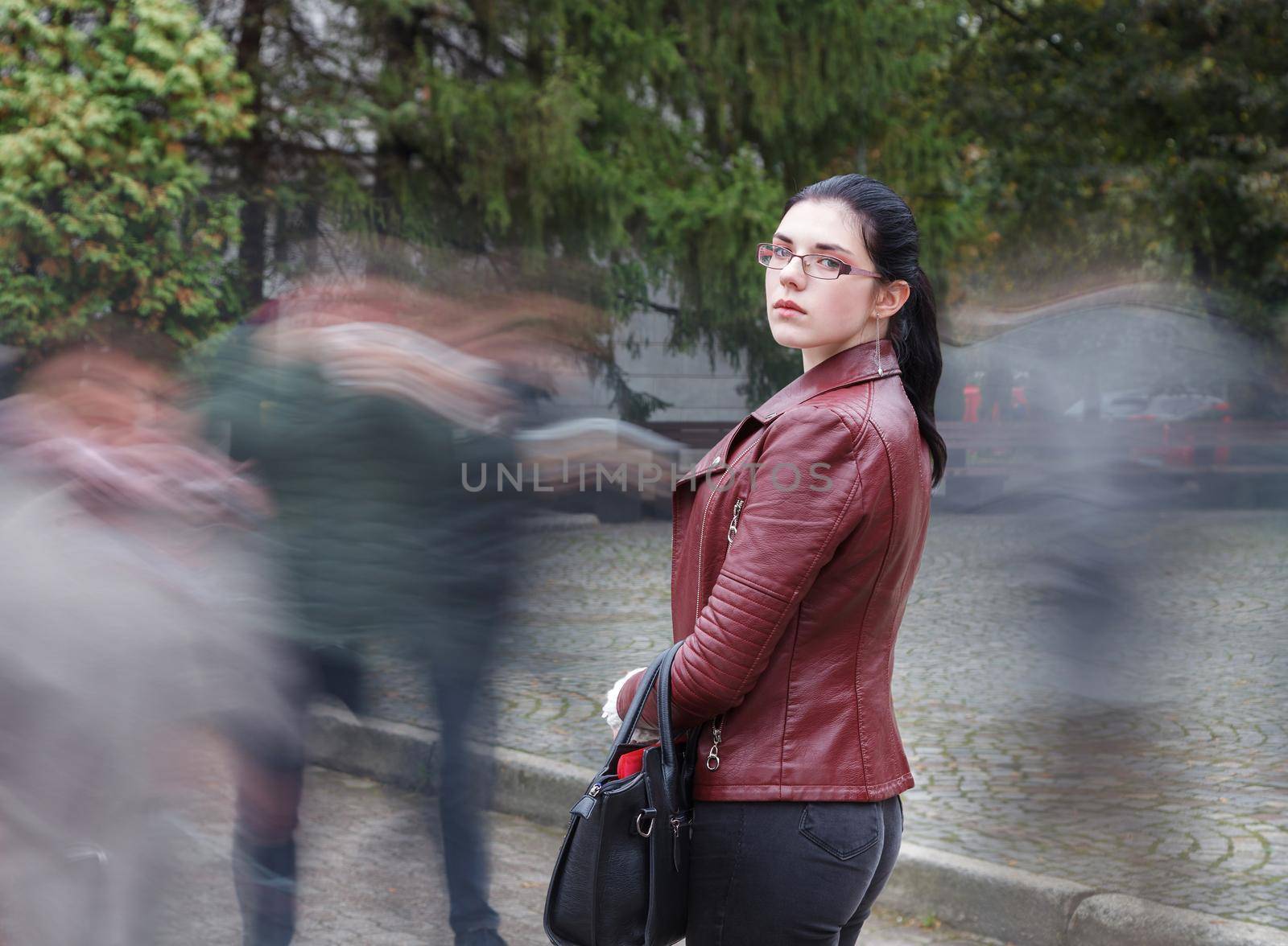 lonely young girl in a brown jacket and black jeans standing on the sidewalk among an indifferent crowd on autumn day