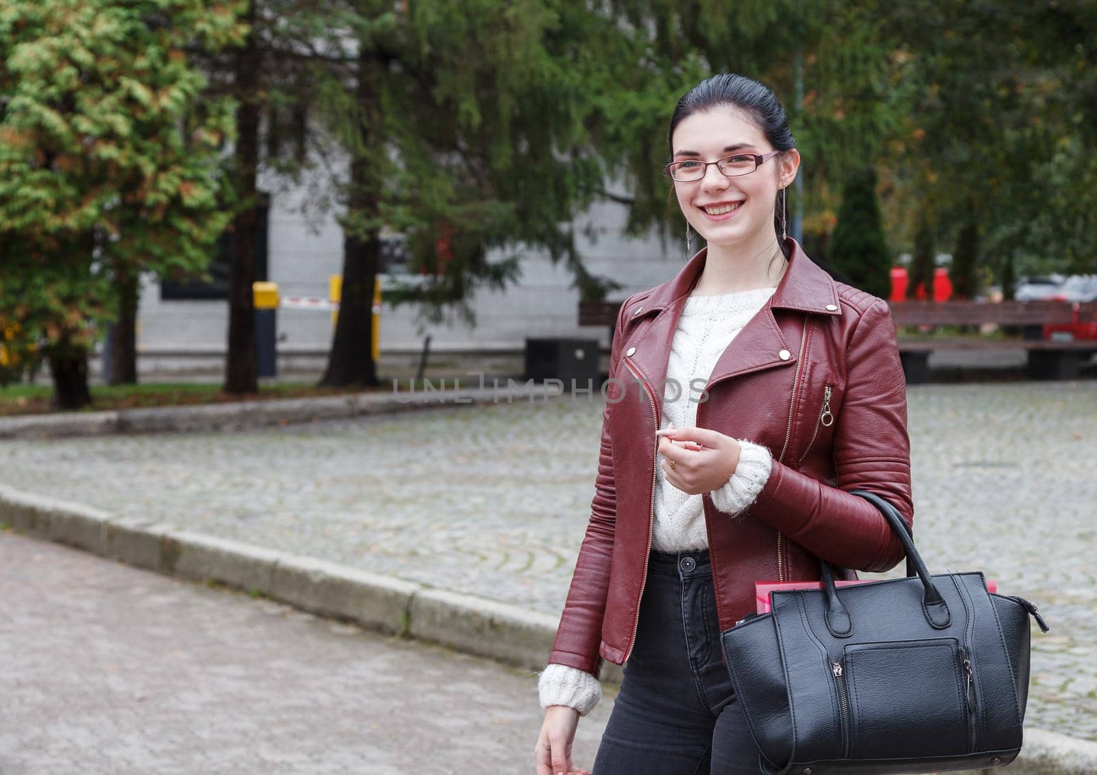 smiling young girl in a brown jacket and black jeans standing on the sidewalk on autumn day