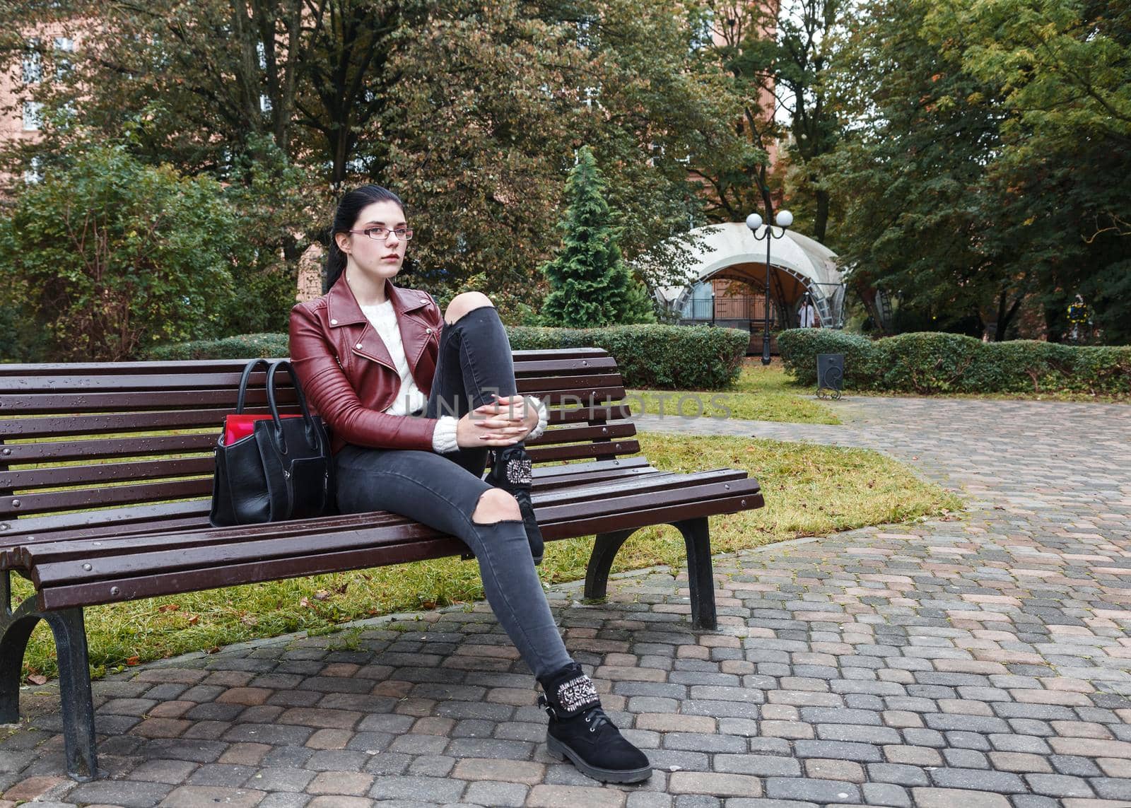 young girl in brown jacket and black jeans sitting on a park bench in autumn