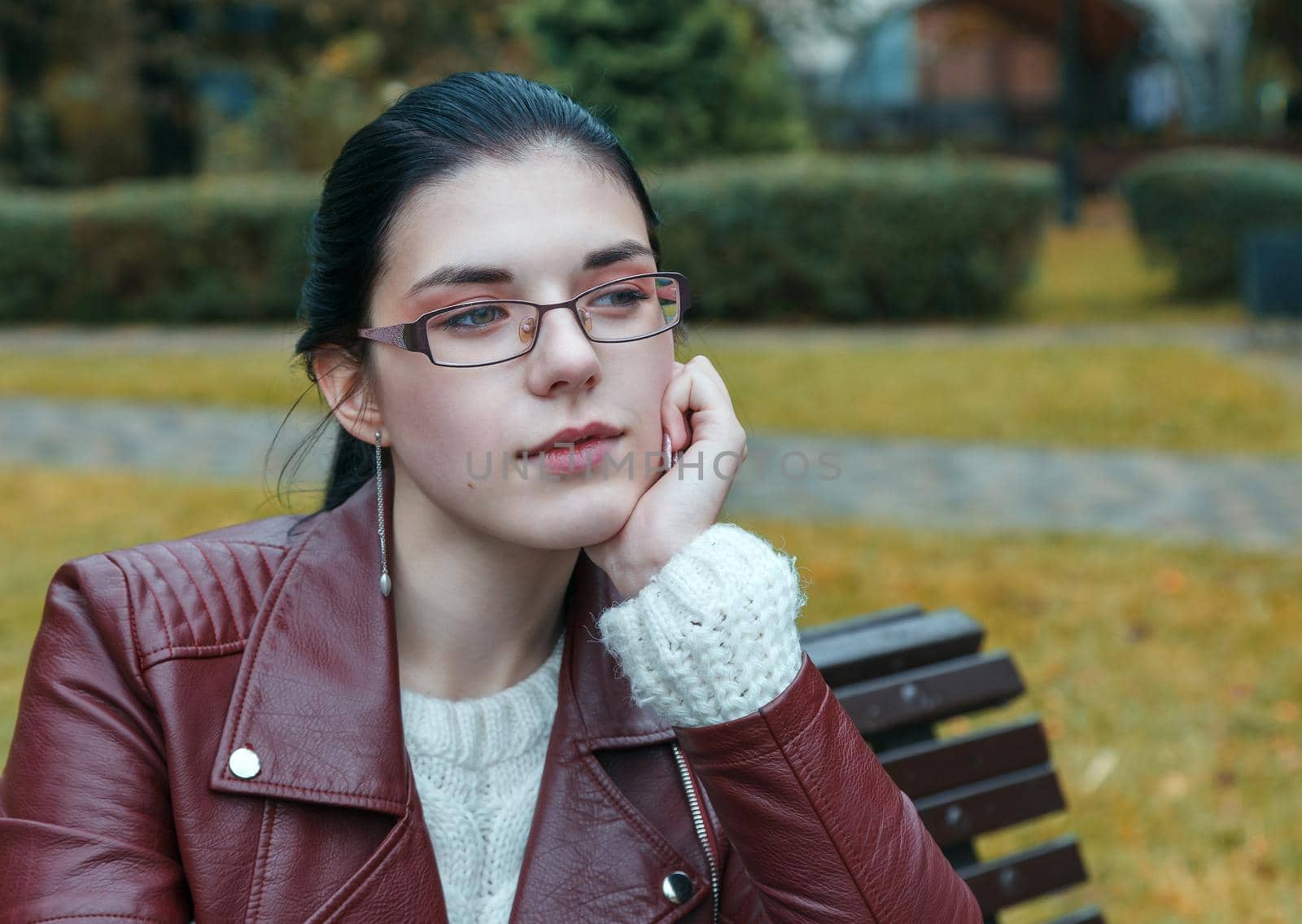 young girl in brown jacket and black jeans sitting on a park bench in autumn. portrait closeup