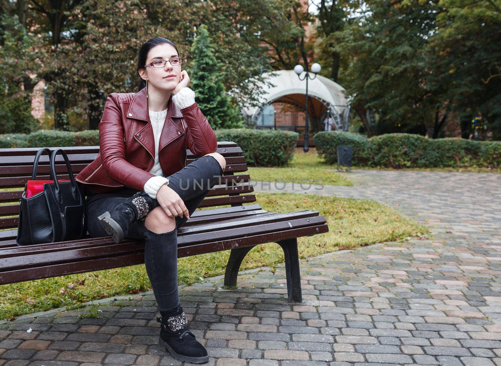 young girl in brown jacket and black jeans sitting on a park bench in autumn