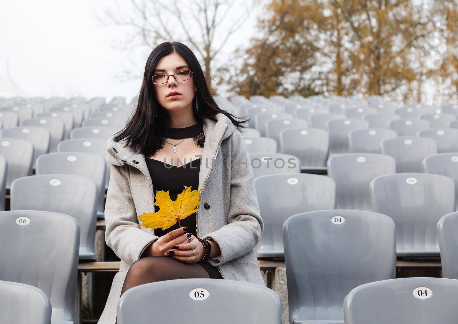 young girl in a gray coat sits on a seat of an empty stadium by raddnatt