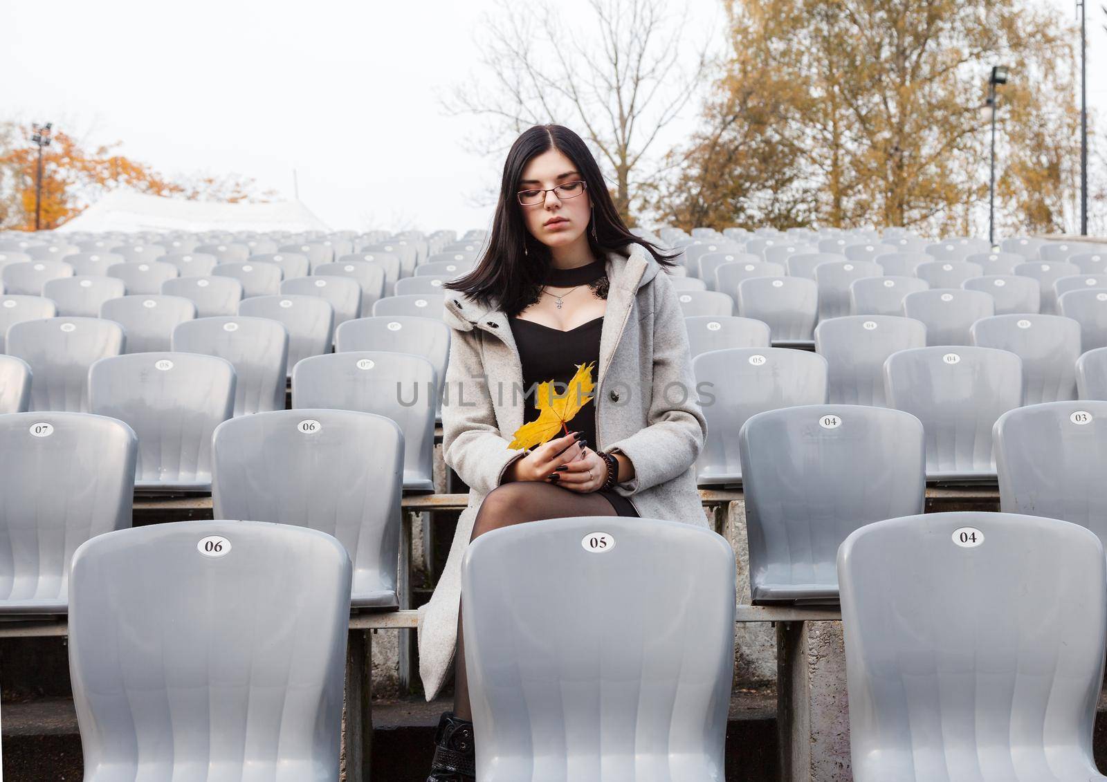 lonely young girl in a gray coat sitting on a seat of an empty stadium on autumn day