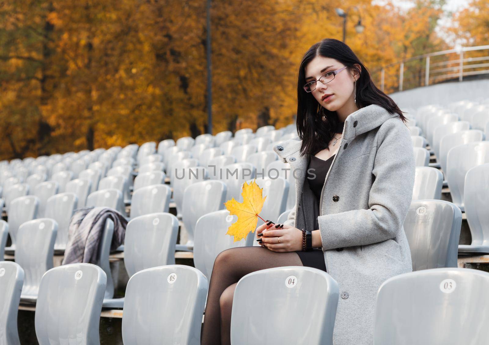 lonely young girl in a gray coat sitting on a seat of an empty stadium on autumn day