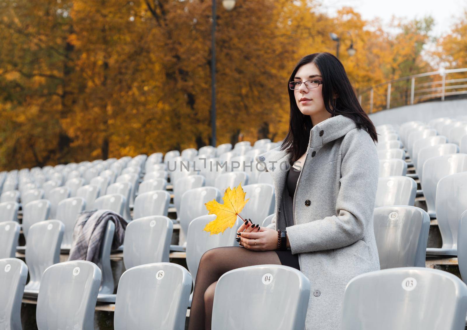 lonely young girl in a gray coat sitting on a seat of an empty stadium on autumn day