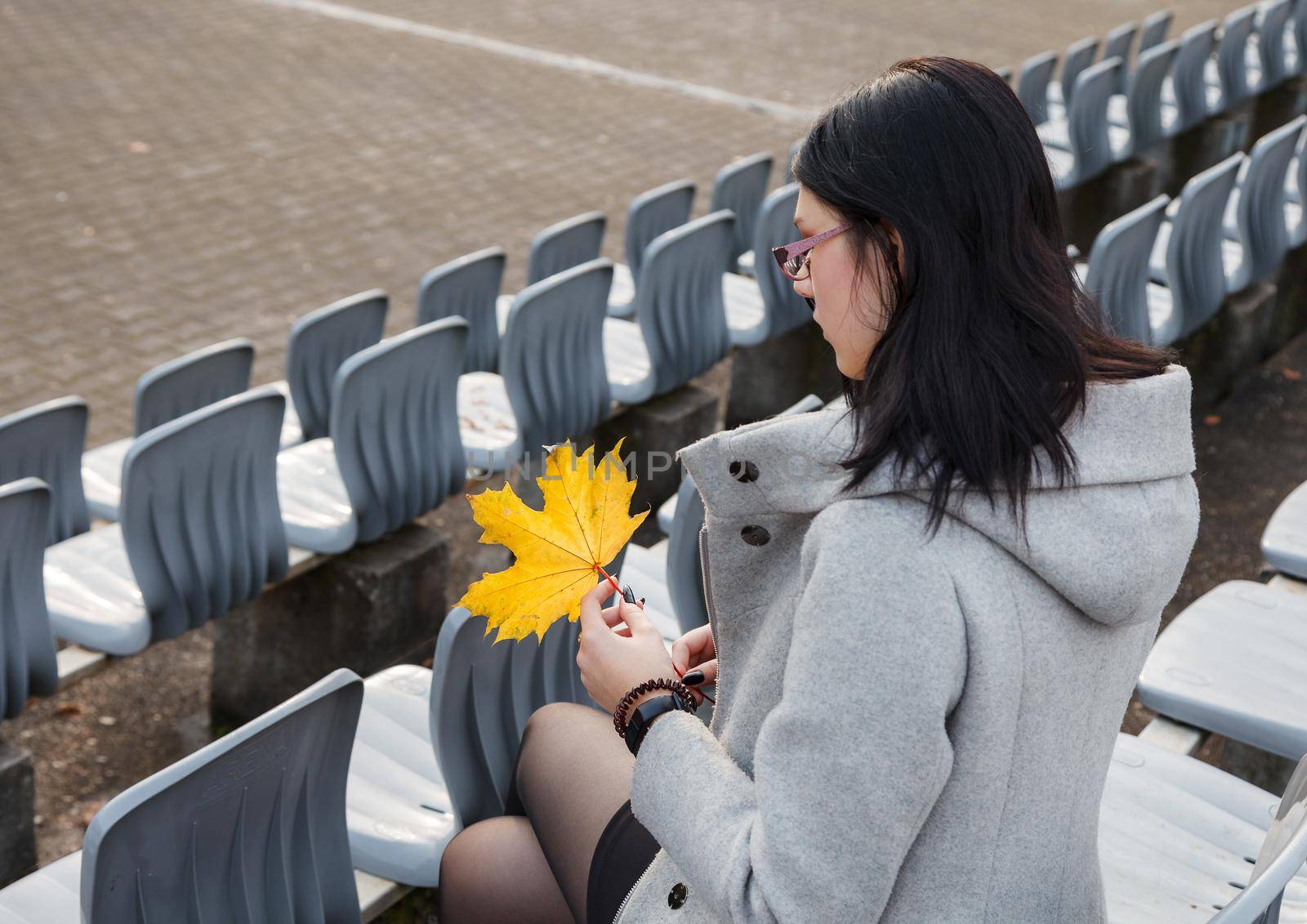 lonely young girl in a gray coat sitting on a seat of an empty stadium on autumn day