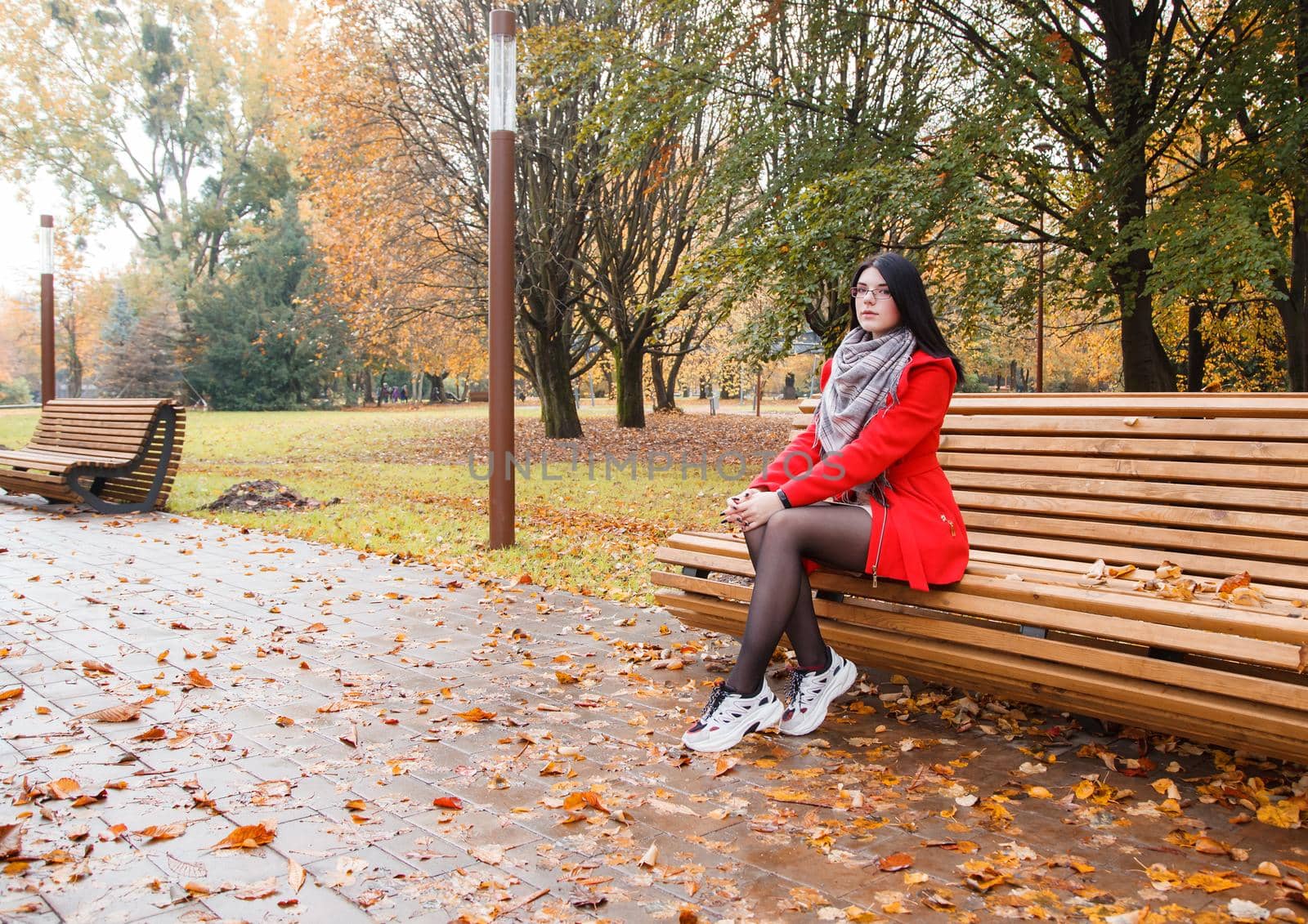 young beautiful girl in a red coat sitting on a bench in a city park after the rain on an autumn day