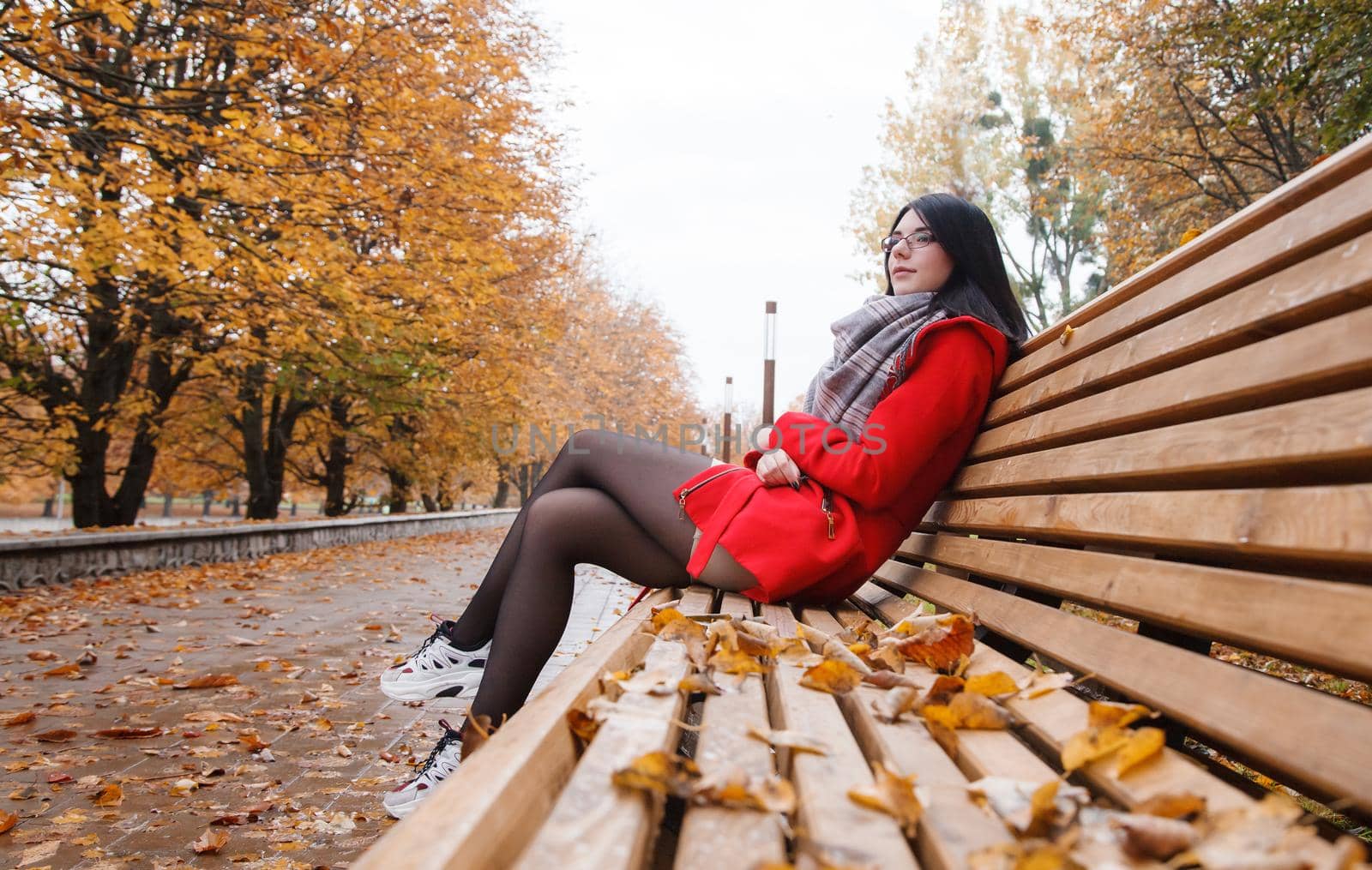 young beautiful girl in a red coat sitting on a bench in a city park after the rain on an autumn day