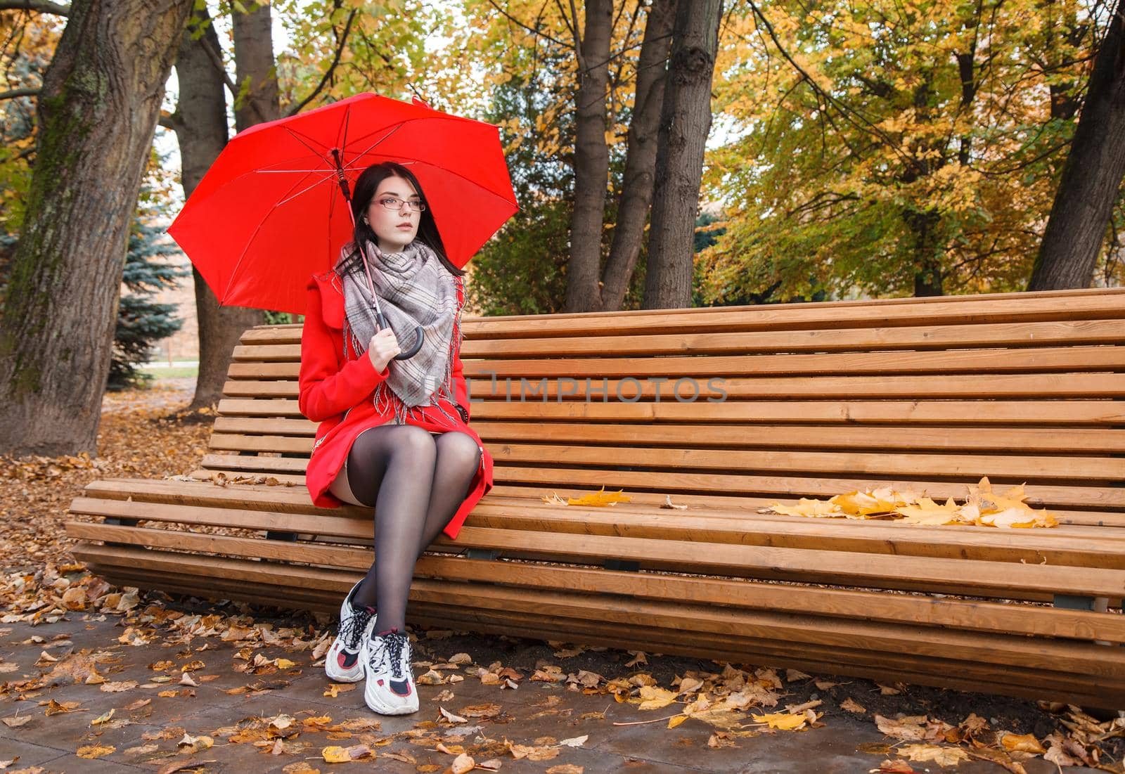 young girl in a red coat with an umbrella sitting on a bench in a city park after the rain on an autumn day