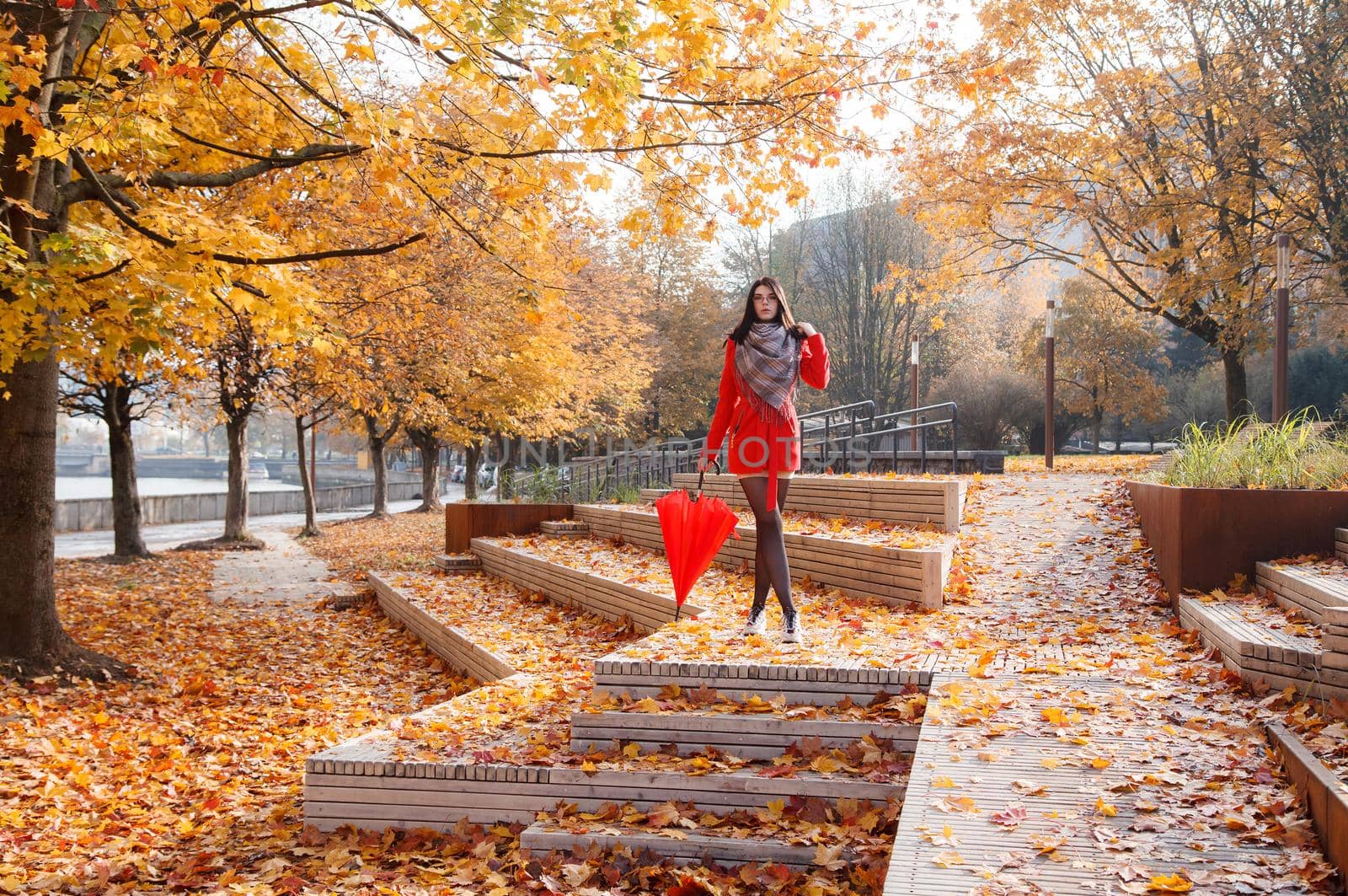 young girl in a red coat with an umbrella standing on the alley of a city park on a sunny autumn day