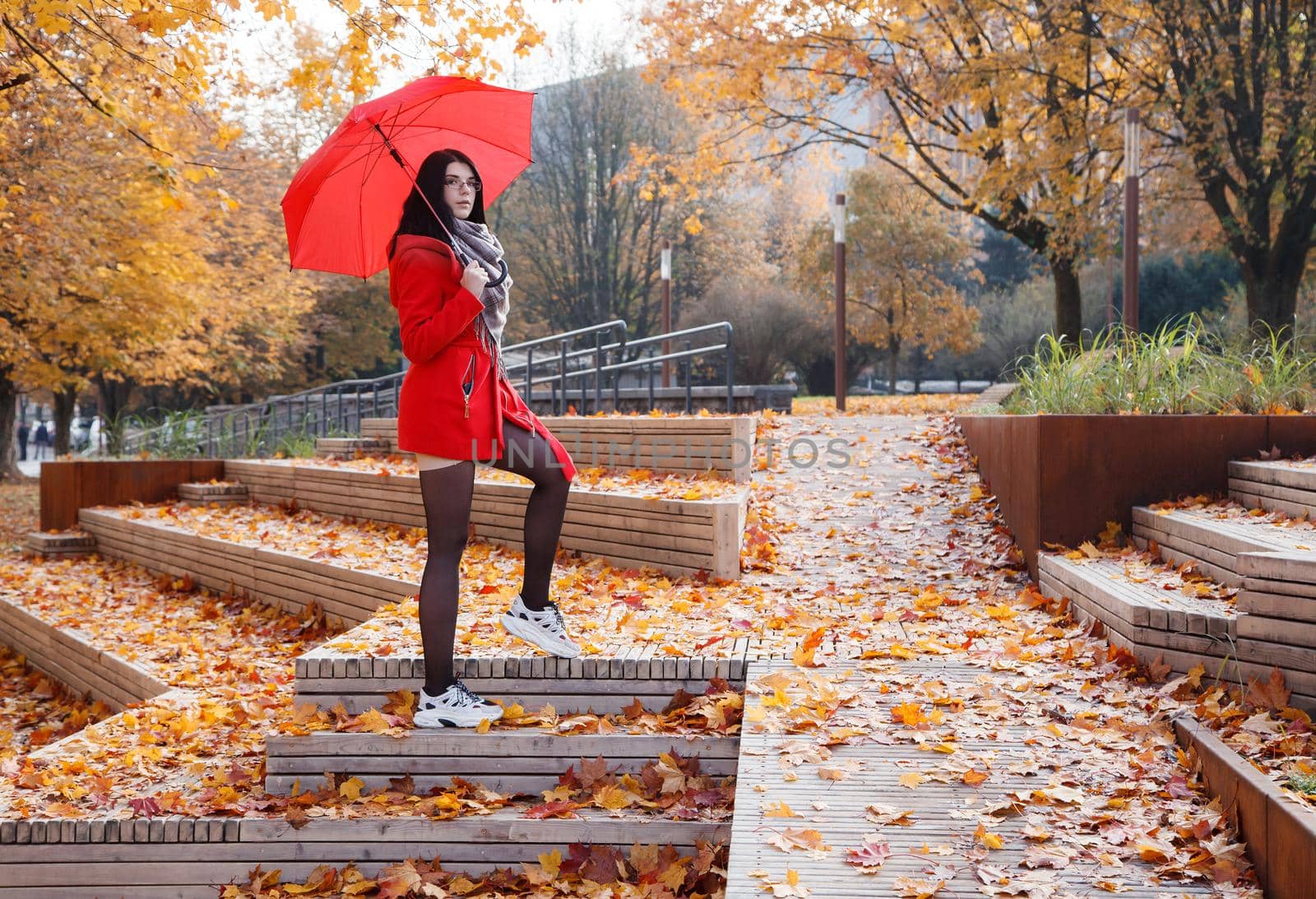 young girl in a red coat with an umbrella standing on the alley of a city park on a sunny autumn day