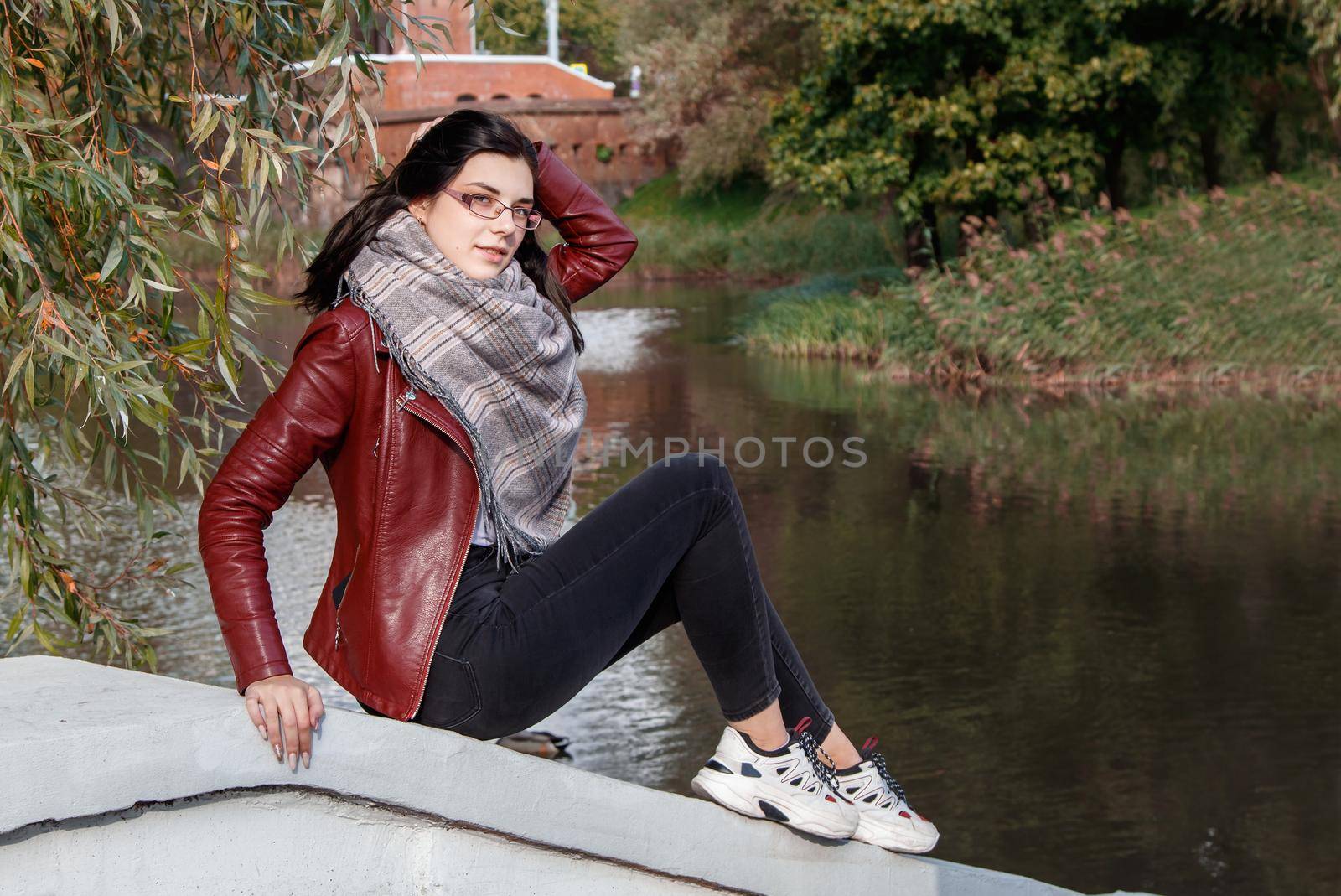 young girl in brown jacket and black jeans sitting on a parapet near a pond in city park on sunny autumn day