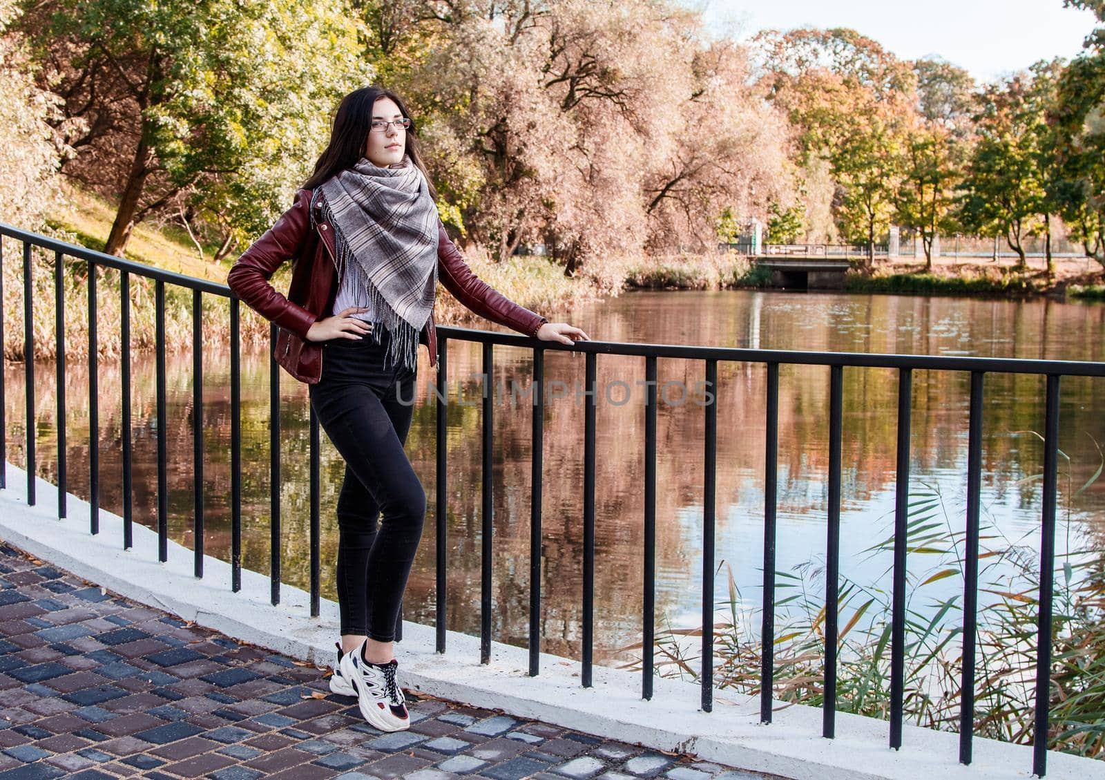 young girl in brown jacket and black jeans standing on bridge near the river in city park on sunny autumn day