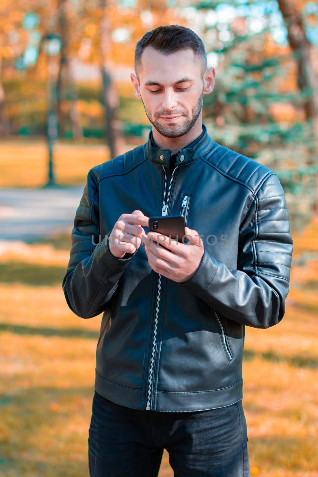 Youthful Guy Using Black Smartphone at the Beautiful Autumn Park. Handsome Young Man with Mobile Phone at Sunny Day - Medium Long Shot