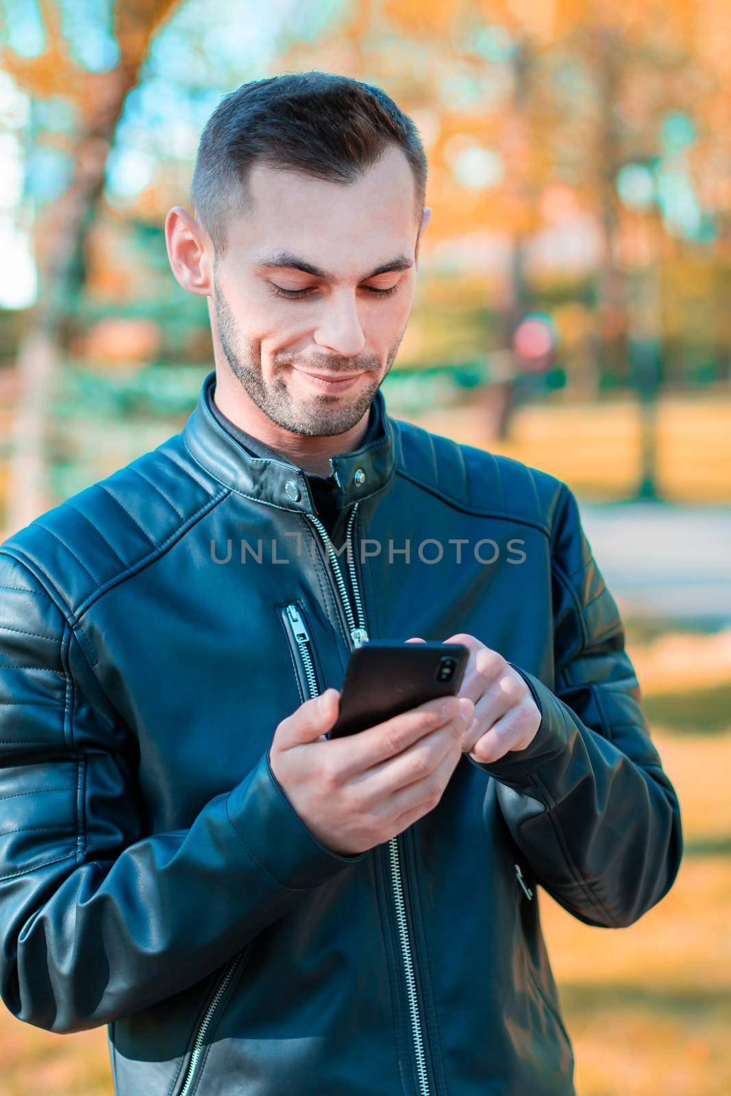 Youthful Guy Using Black Smartphone at the Beautiful Autumn Park. Handsome Young Man with Mobile Phone at Sunny Day - Medium Shot Portrait