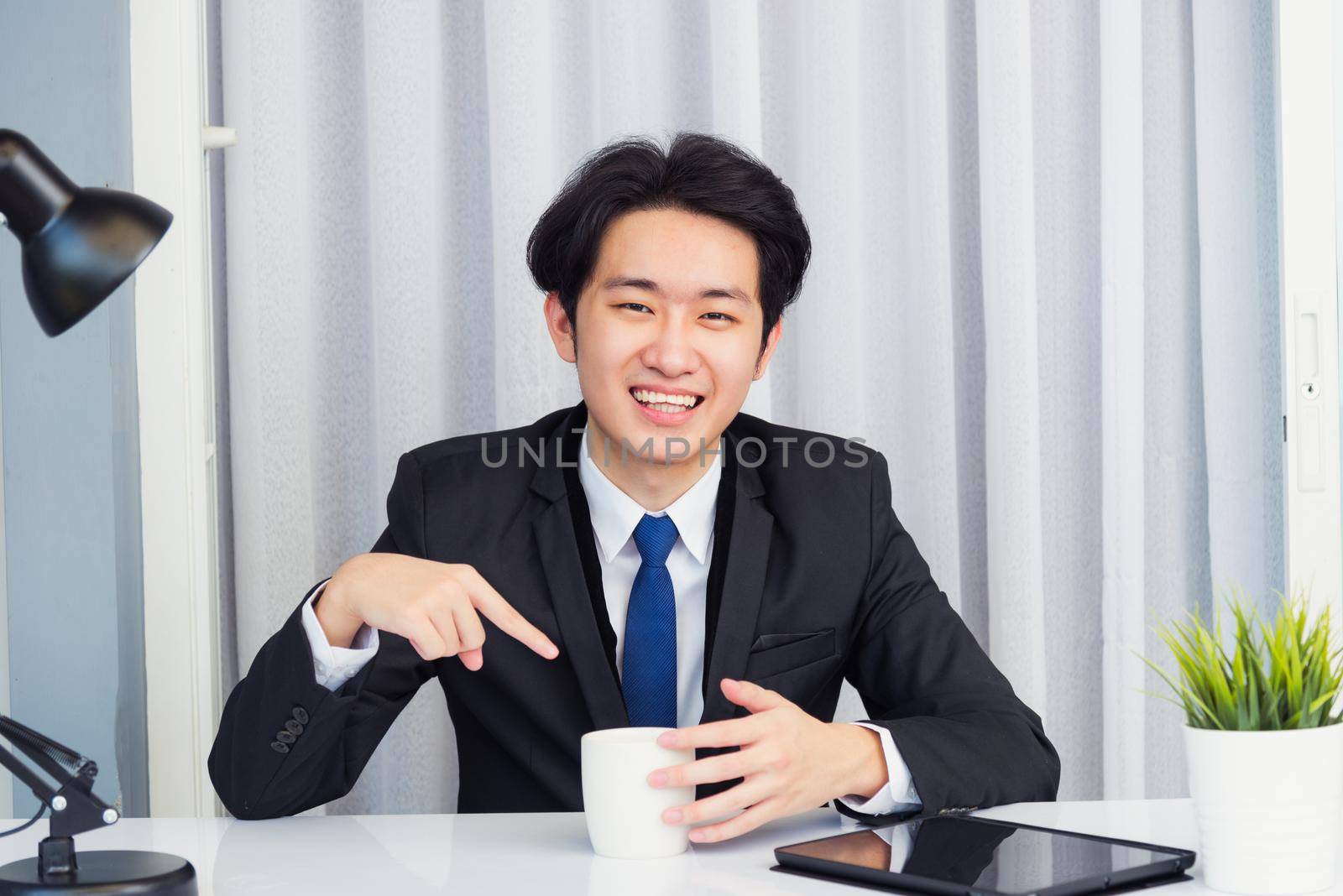 Work from home, Asian young businessman video conference call or facetime he looking to camera sitting on desk point coffee cup to invite colleagues drink coffee before event at home office