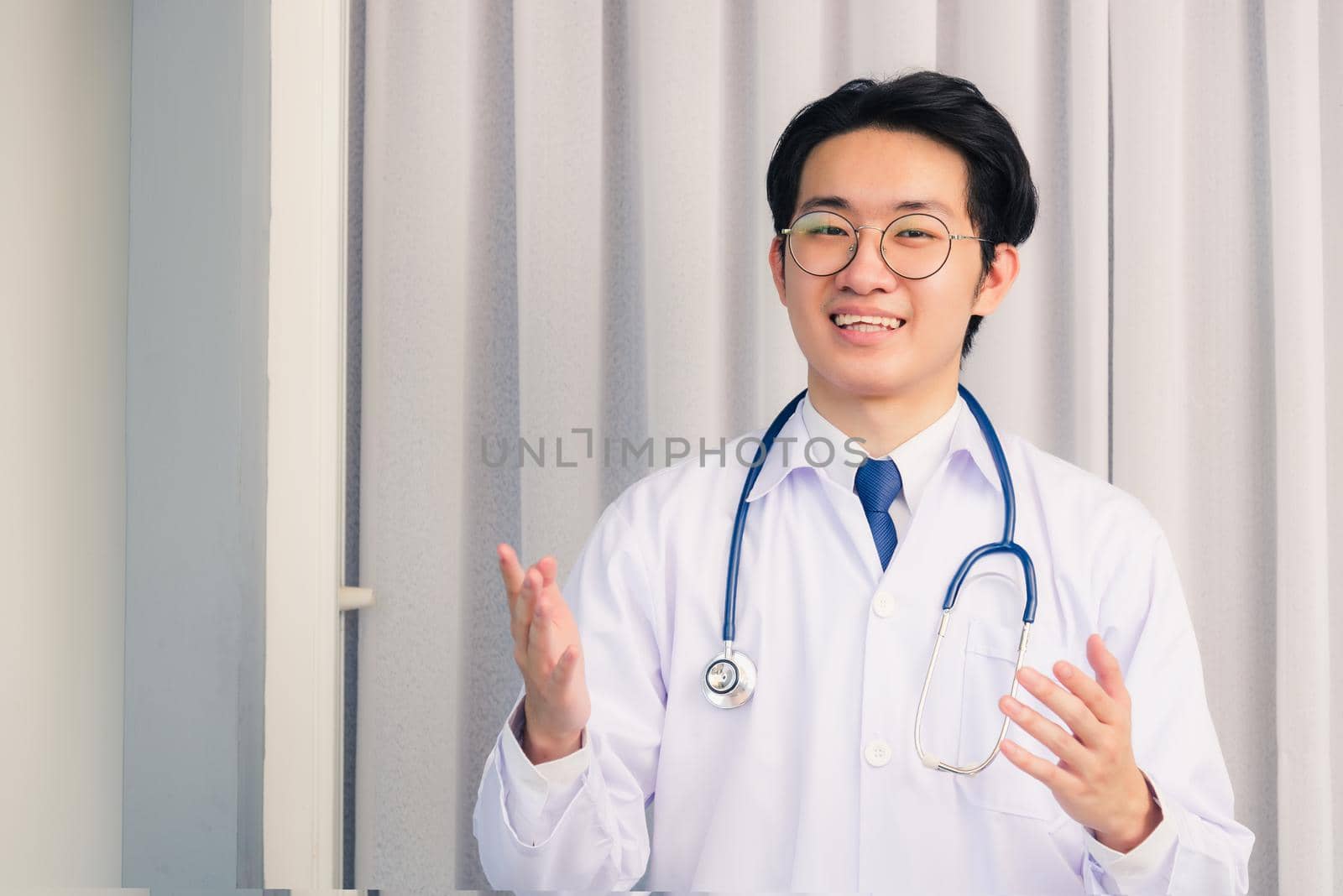 Portrait of Happy Asian young doctor handsome man smiling in uniform with stethoscope talking online video conference call or facetime raise his hand to explain, healthcare medicine concept