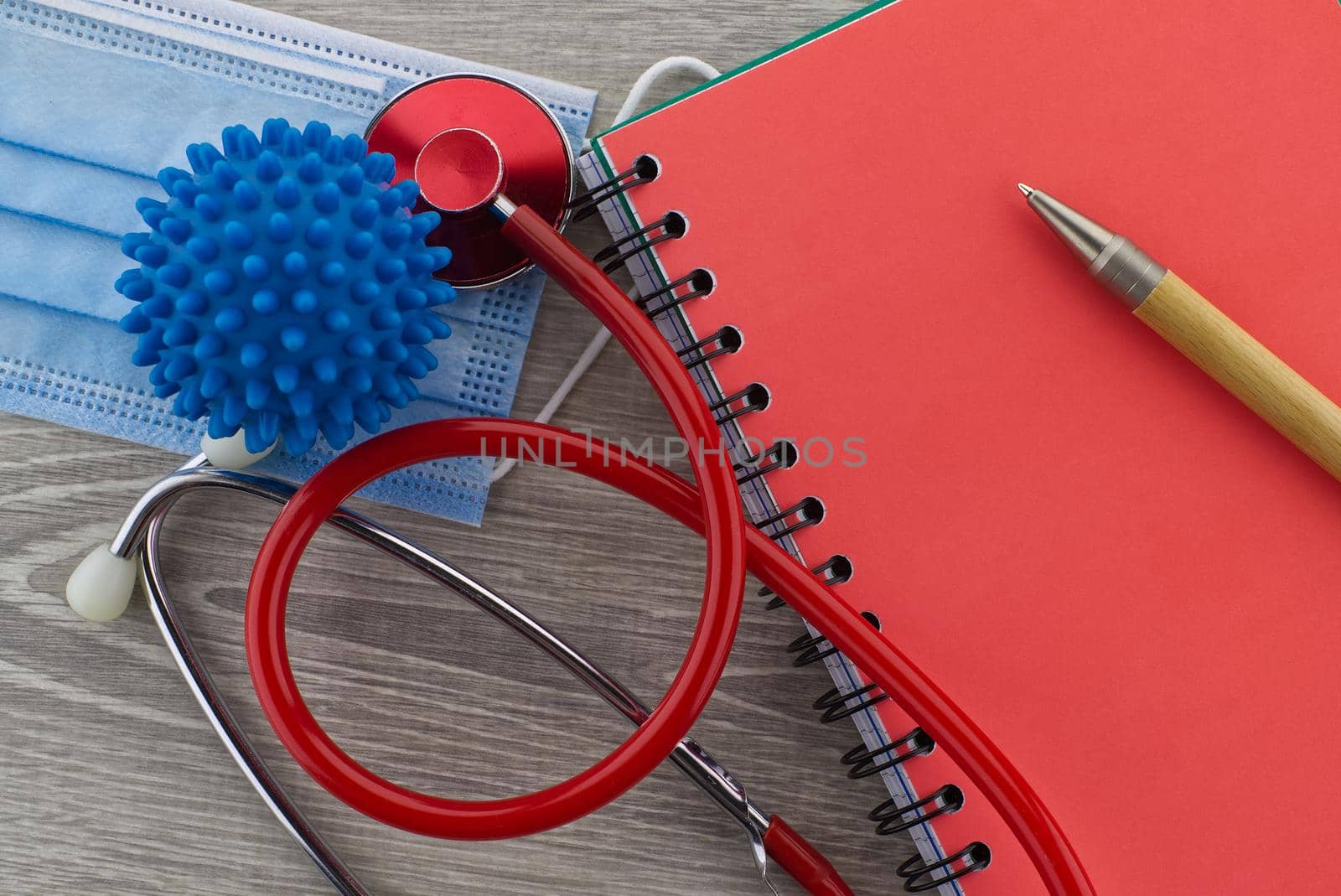 Virus pandemic and medical themed still life with a colorful red stethoscope and surgical face mask placed to the side of wire bound notepad and pen viewed as a flat lay from overhead