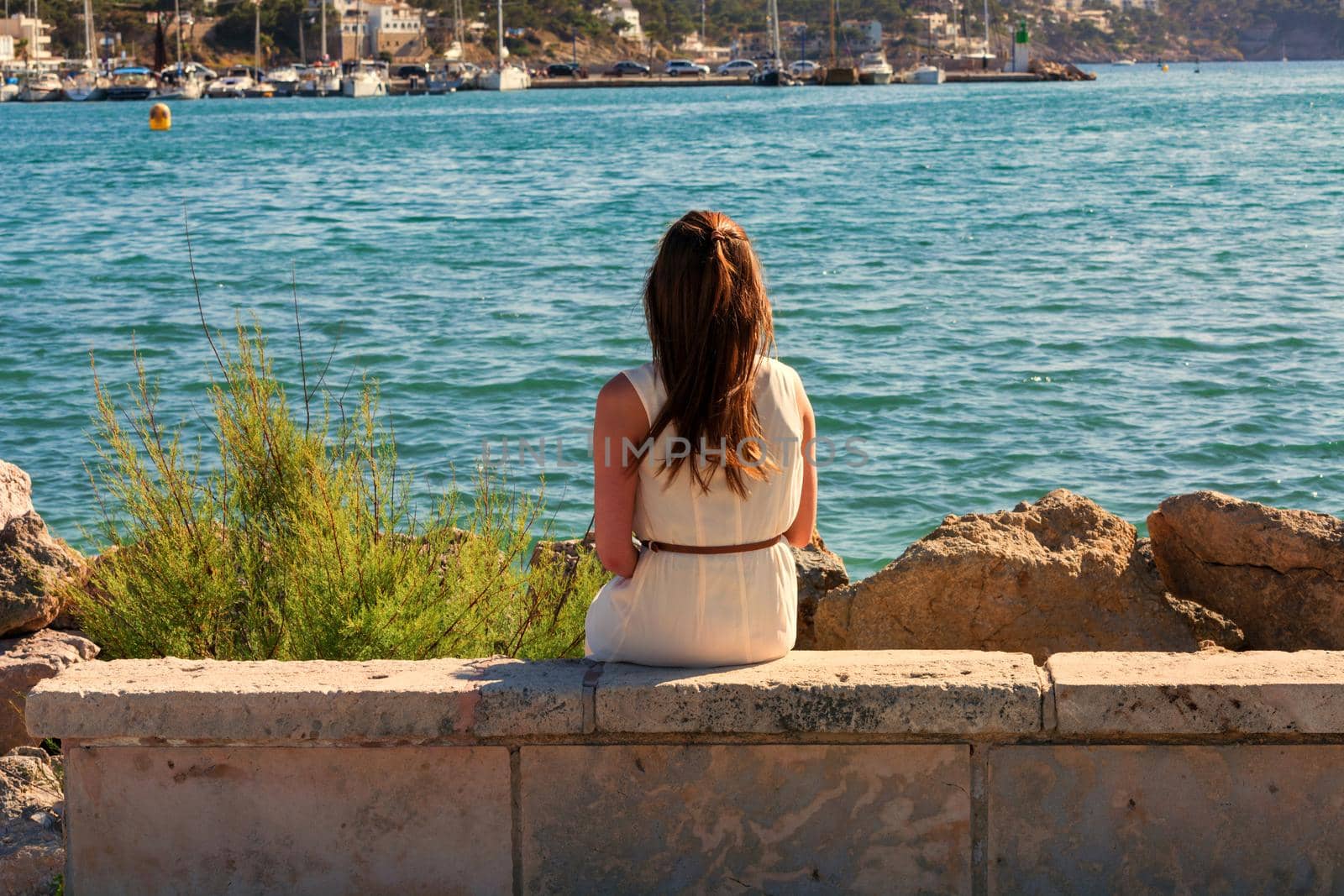 Young attractive woman sitting on a wall at the Mediterranean Sea looking at the harbor.