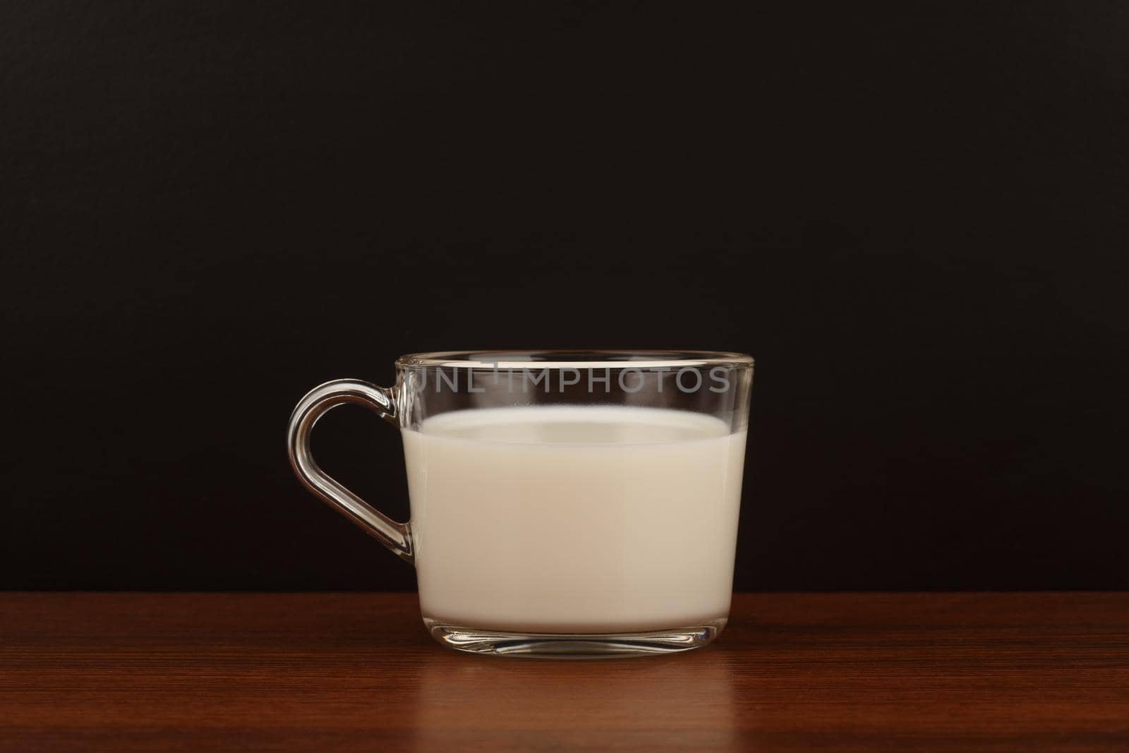 Transparent cup of milk on brown wooden table against black background with copy space by Senorina_Irina