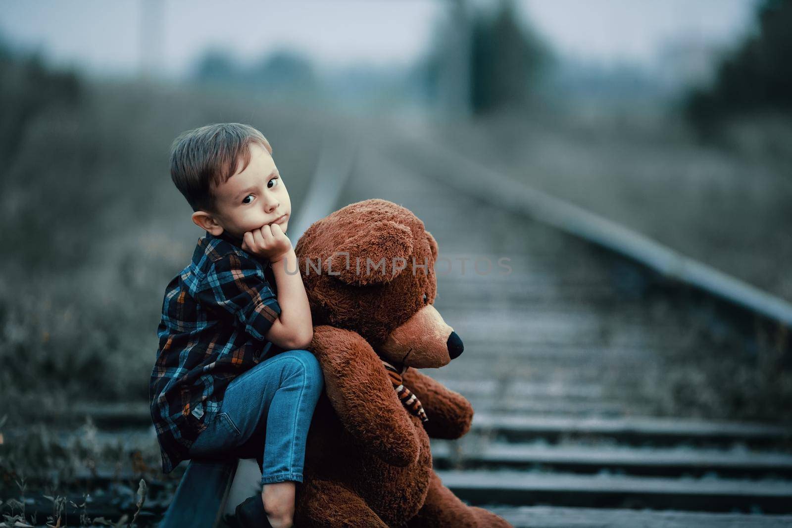 An abandoned homeless child, an orphan. A lonely boy hugs a stuffed toy and sits on the tracks, looking sadly at the camera. Street children from dysfunctional families.