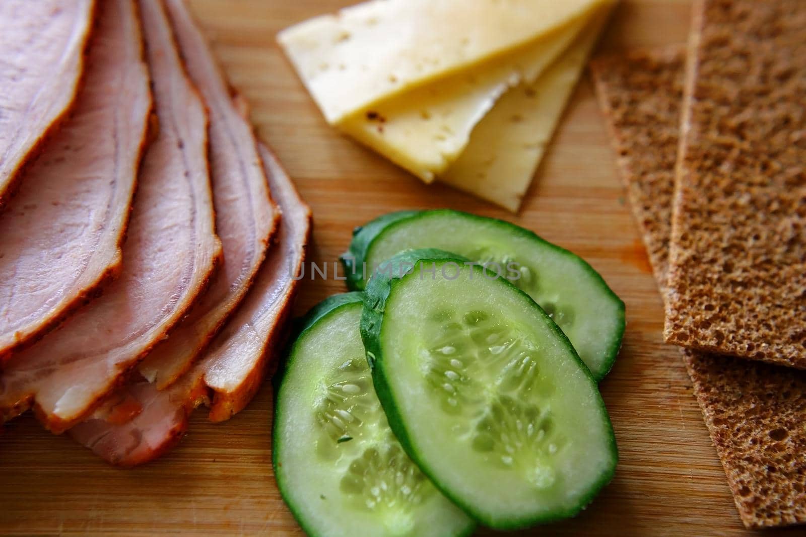 Slicing ham, cucumbers and cheese with ruddy thin breads lie on a wooden board. A healthy and healthy breakfast. A close-up of the top.