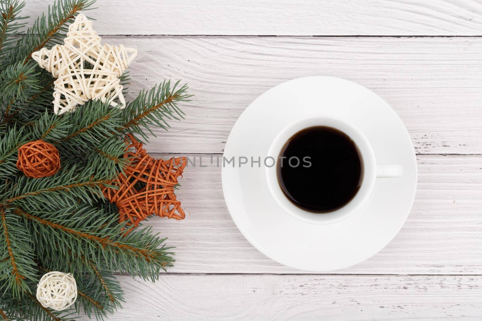 Flat lay with white ceramic coffee cup with a saucer on white wooden table decorated with Christmas tree and rotang stars with balls