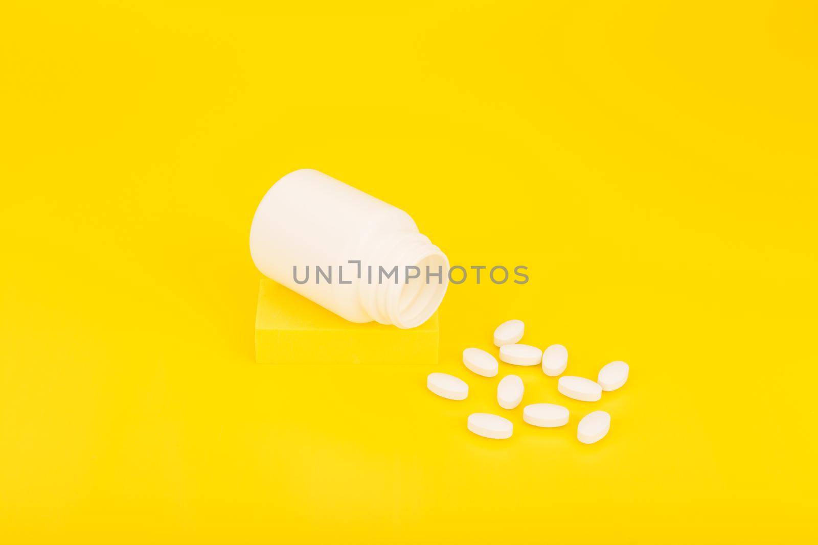 Still life with opened white medication bottle with spilled oval pills on podium against yellow background. Minimalistic concept of vitamins, minerals, supplement and healthcare