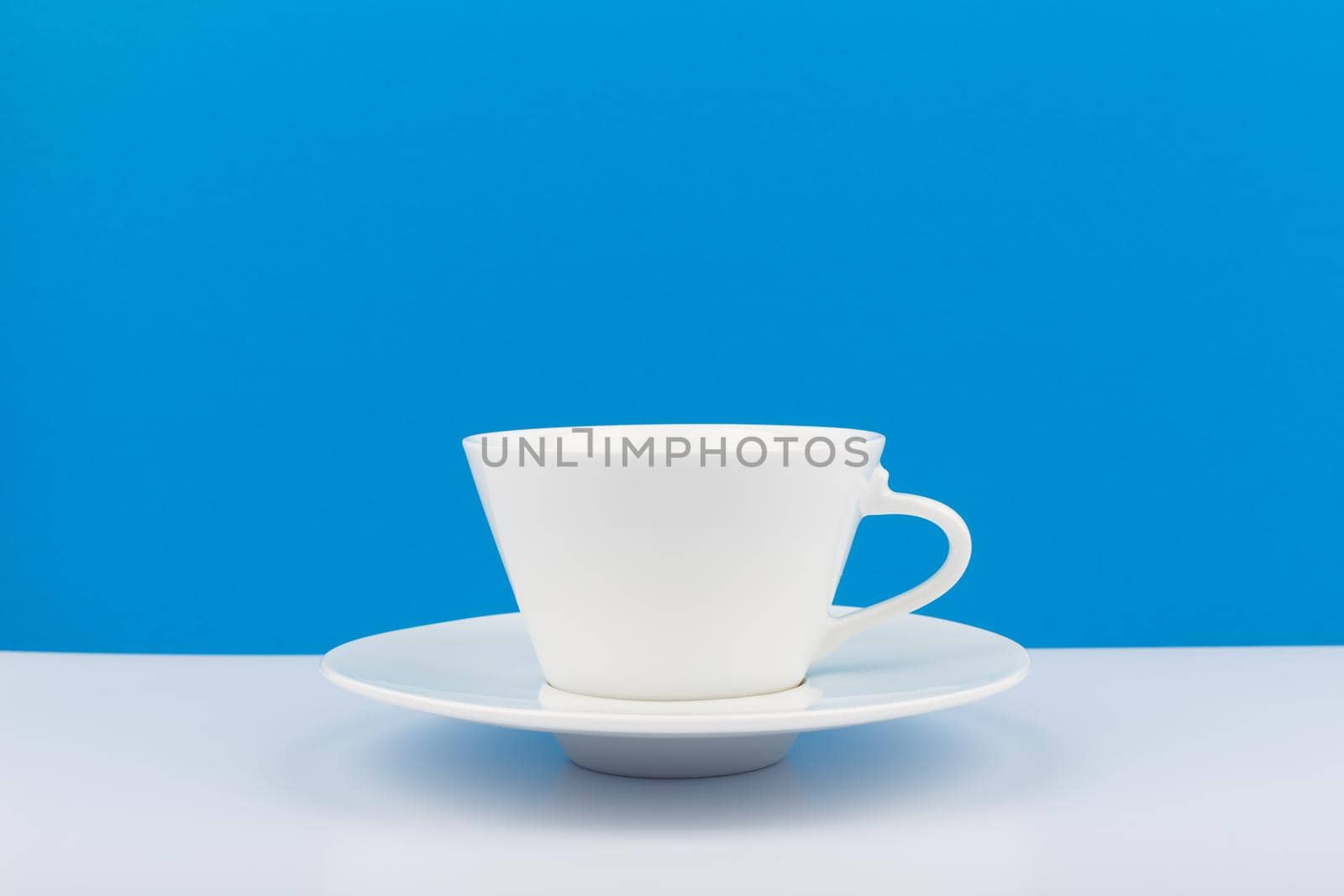 Minimalistic still life with white ceramic coffee cup with a saucer on white table against blue background with space for text