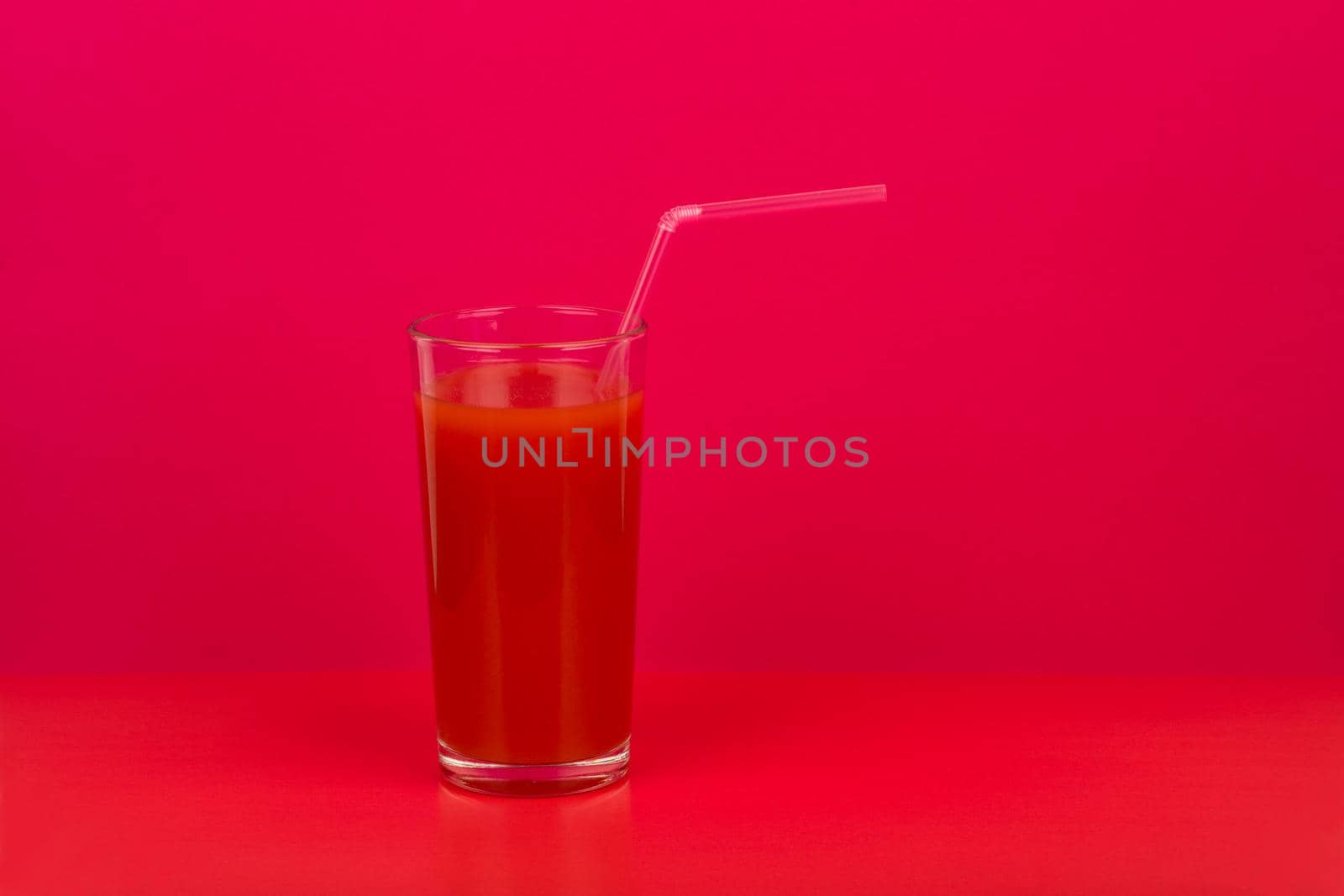 Simple still life with tomato juice in a glass with a straw against red background with a space for text. Creative concept of fresh juices and healthy lifestyle or wellbeing