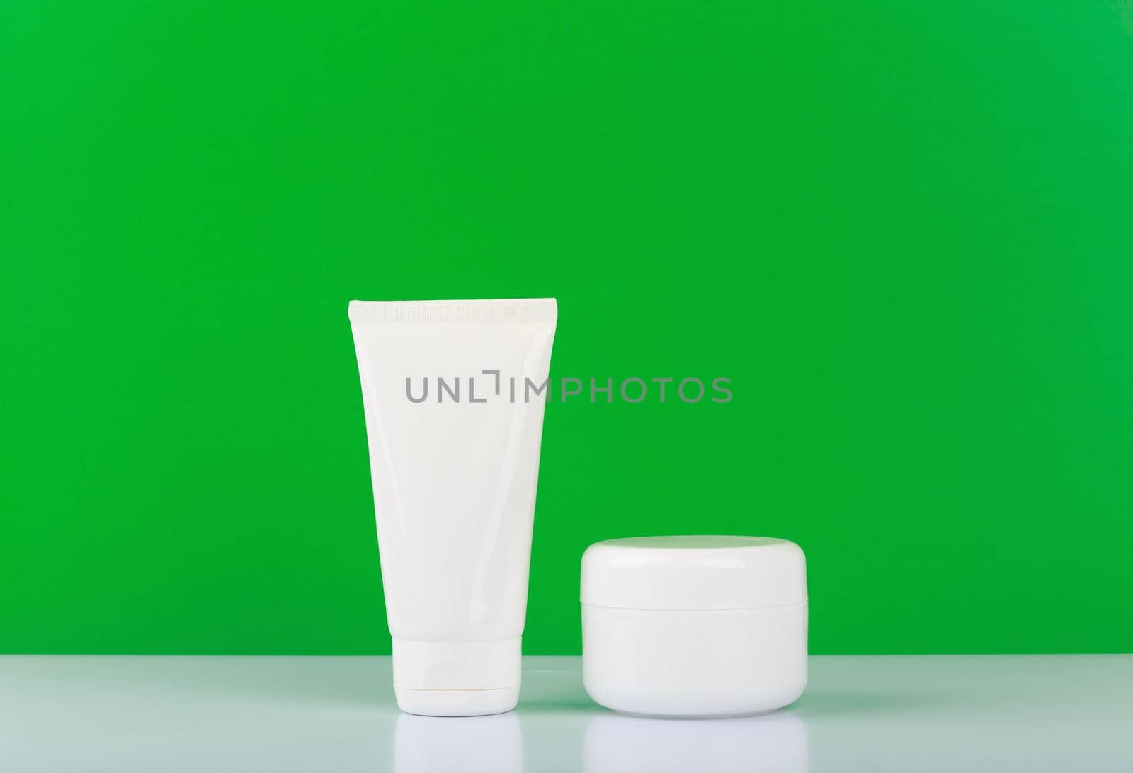 Simple still life with unbranded face and hand cream on white glossy table against green background with space for text. Concept of organic skin cosmetics or cosmetic set for hands and face 