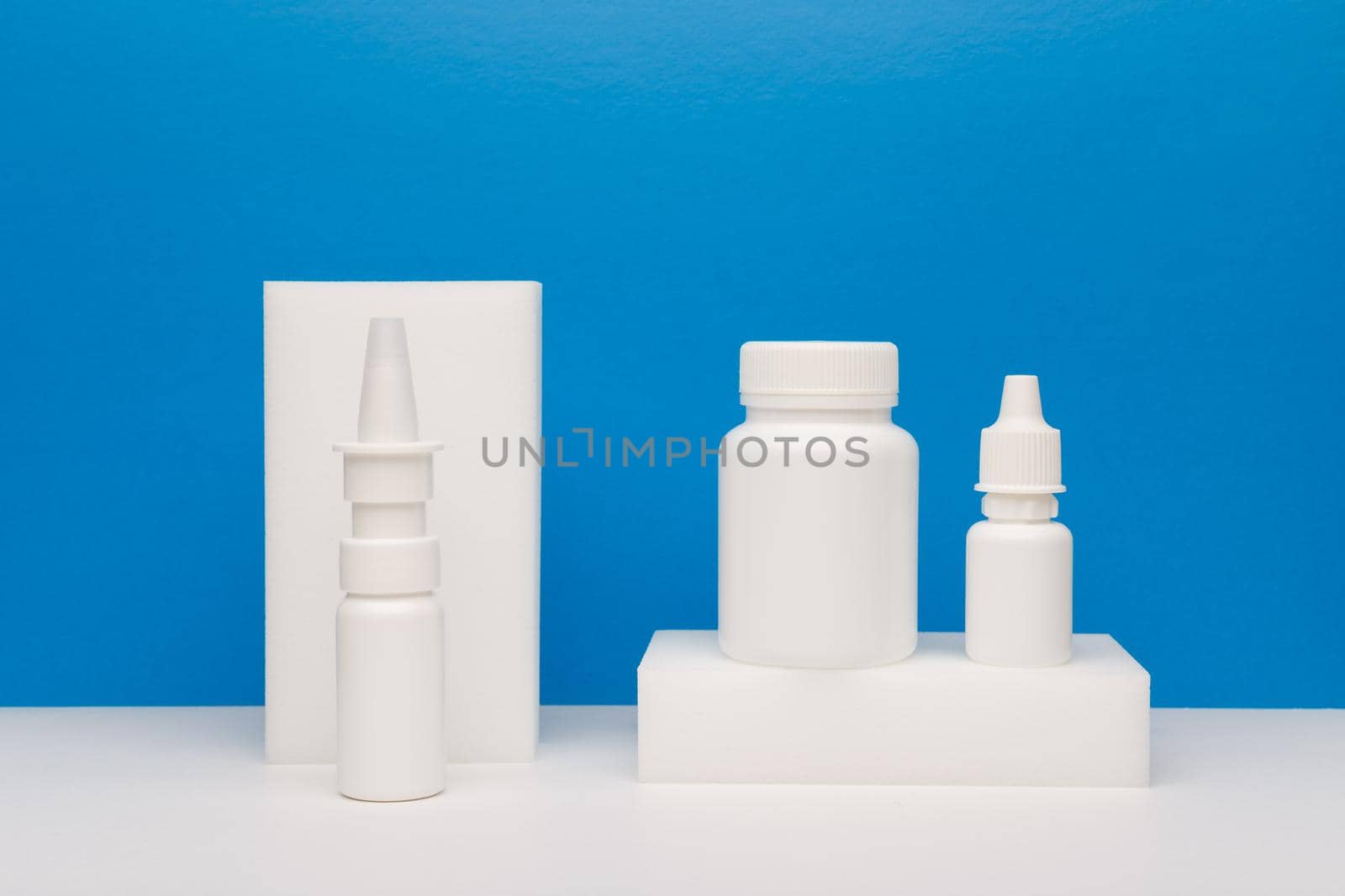 Medical still life with nose spay, eye drops and pills on white podium against blue background by Senorina_Irina