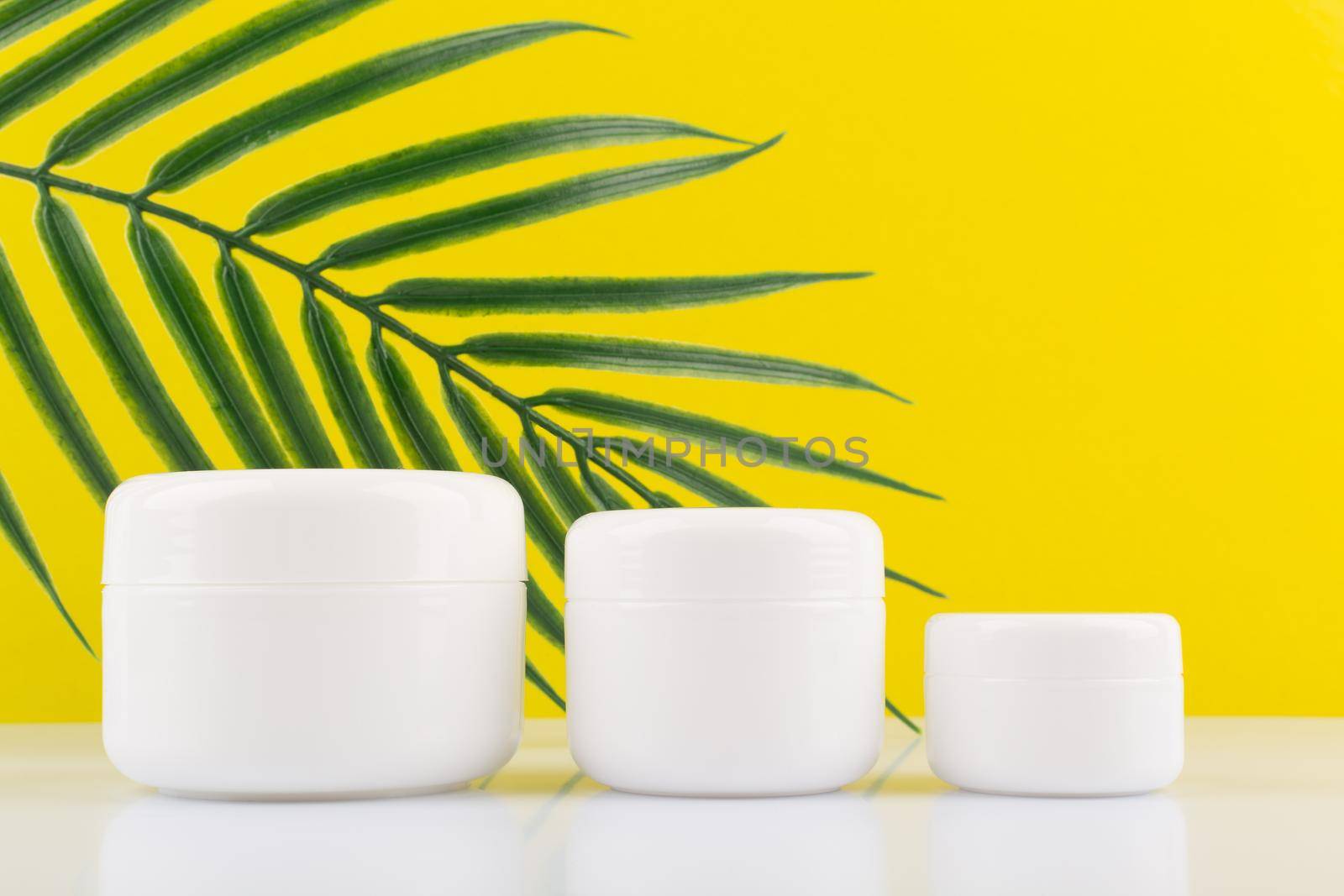 Still life with set of white unbranded glossy cream cream jars on white table against yellow background with palm leaf with a space for text. Concept of organic skin care products . High quality photo
