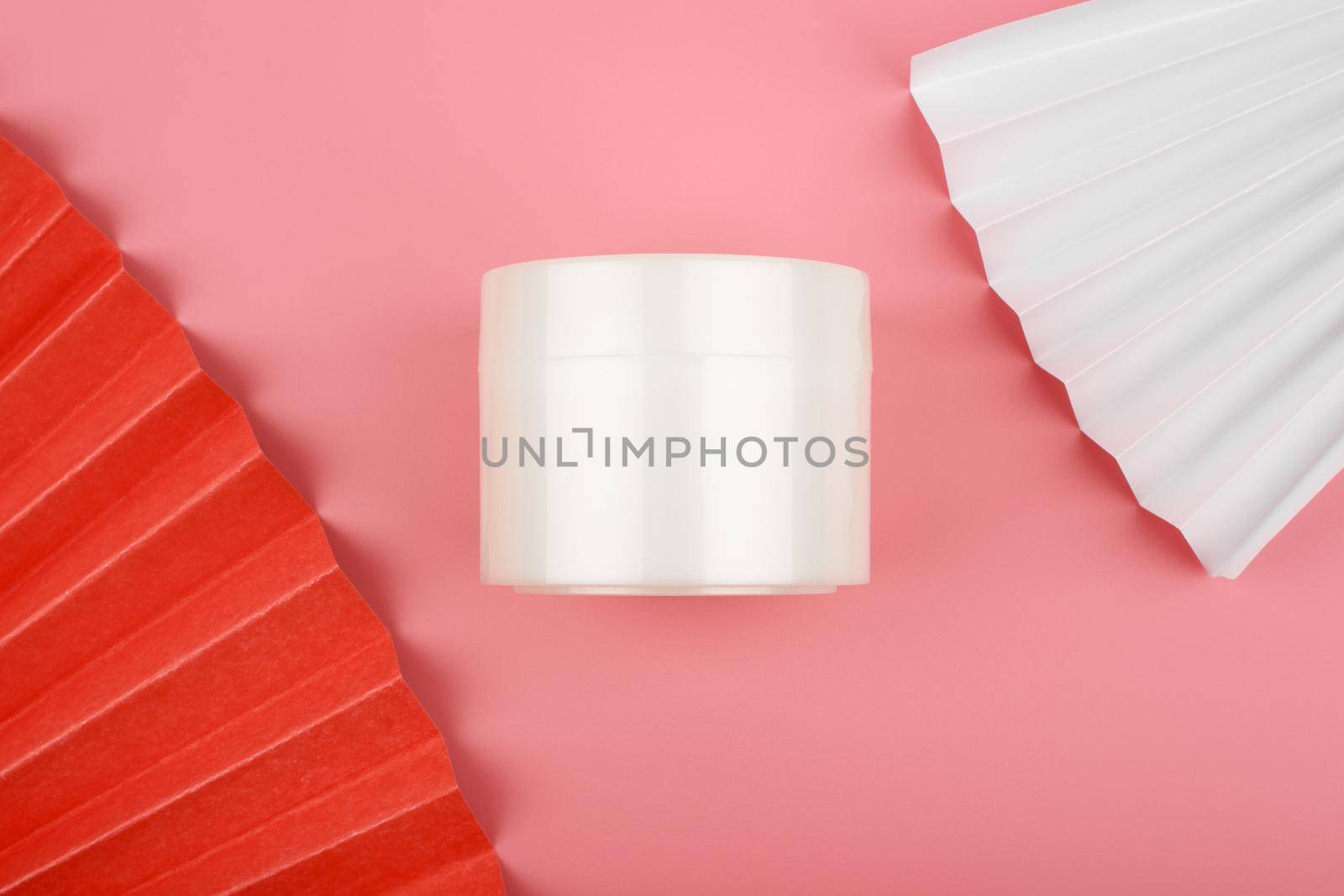 Minimalistic flat lay with white glossy cream jar on pink background decorated with white and red wavers. High quality photo