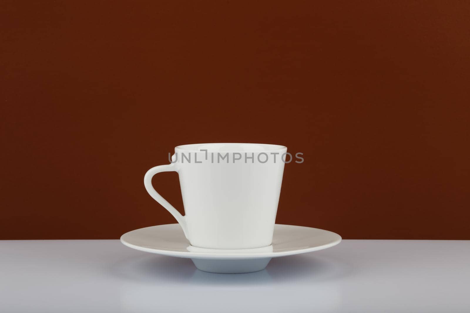 Still life with white ceramic coffee cup on plate against brown background with space for text. 