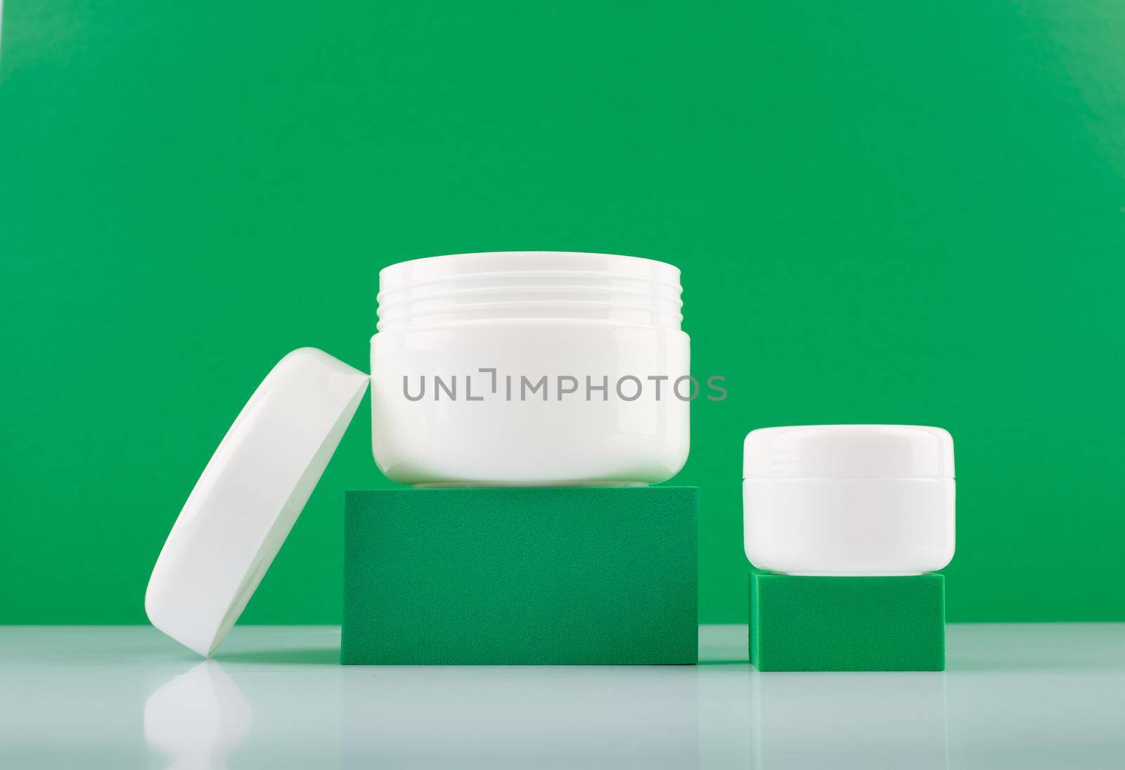 Still life with two white glossy cream jars on podiums against green background with space for text. Concept of beauty, skin care or organic cosmetic with natural ingredients