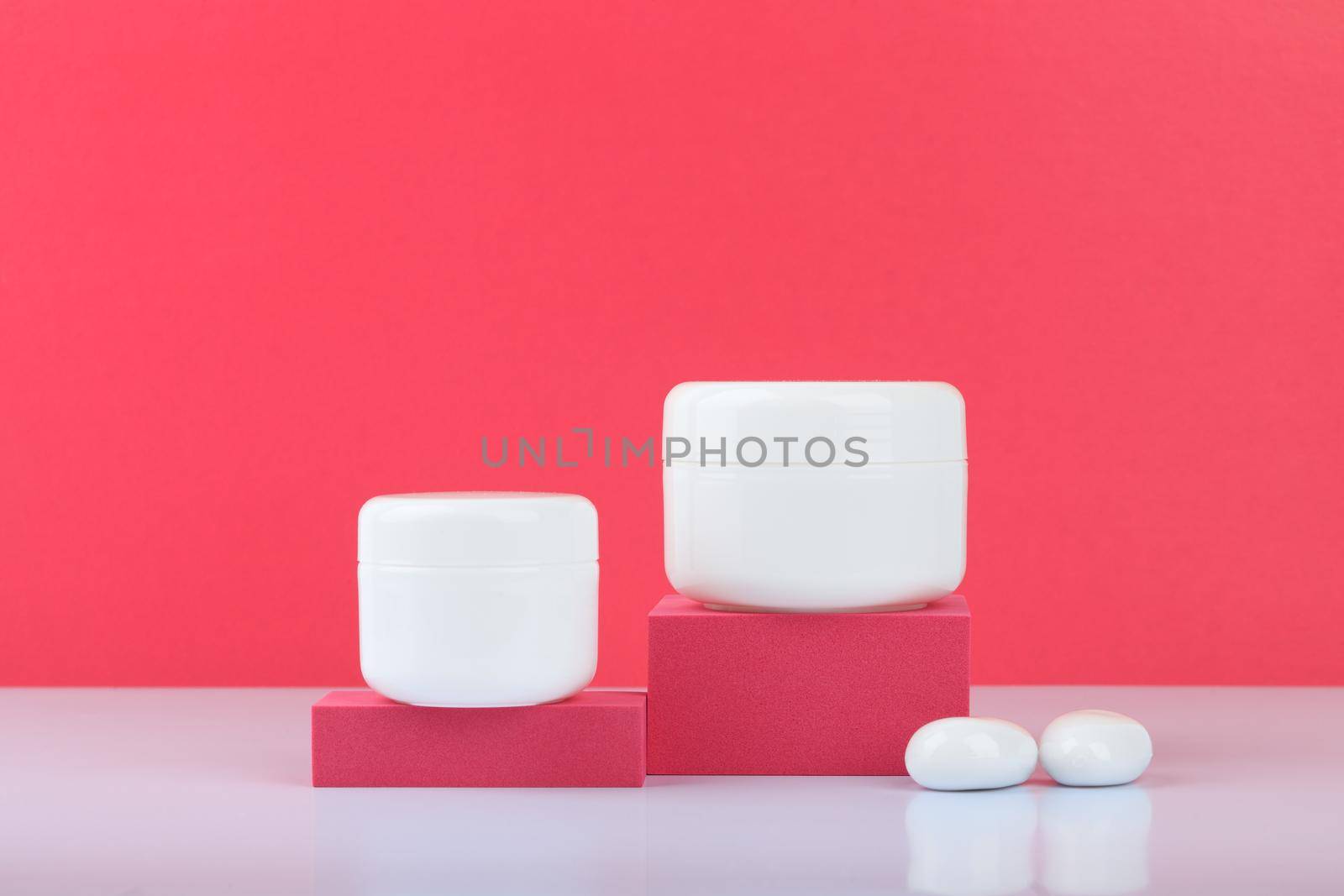 Still life with white glossy cream jars on podiums with white stones on white table against pink background with space for text. Concept of beauty. High quality photo